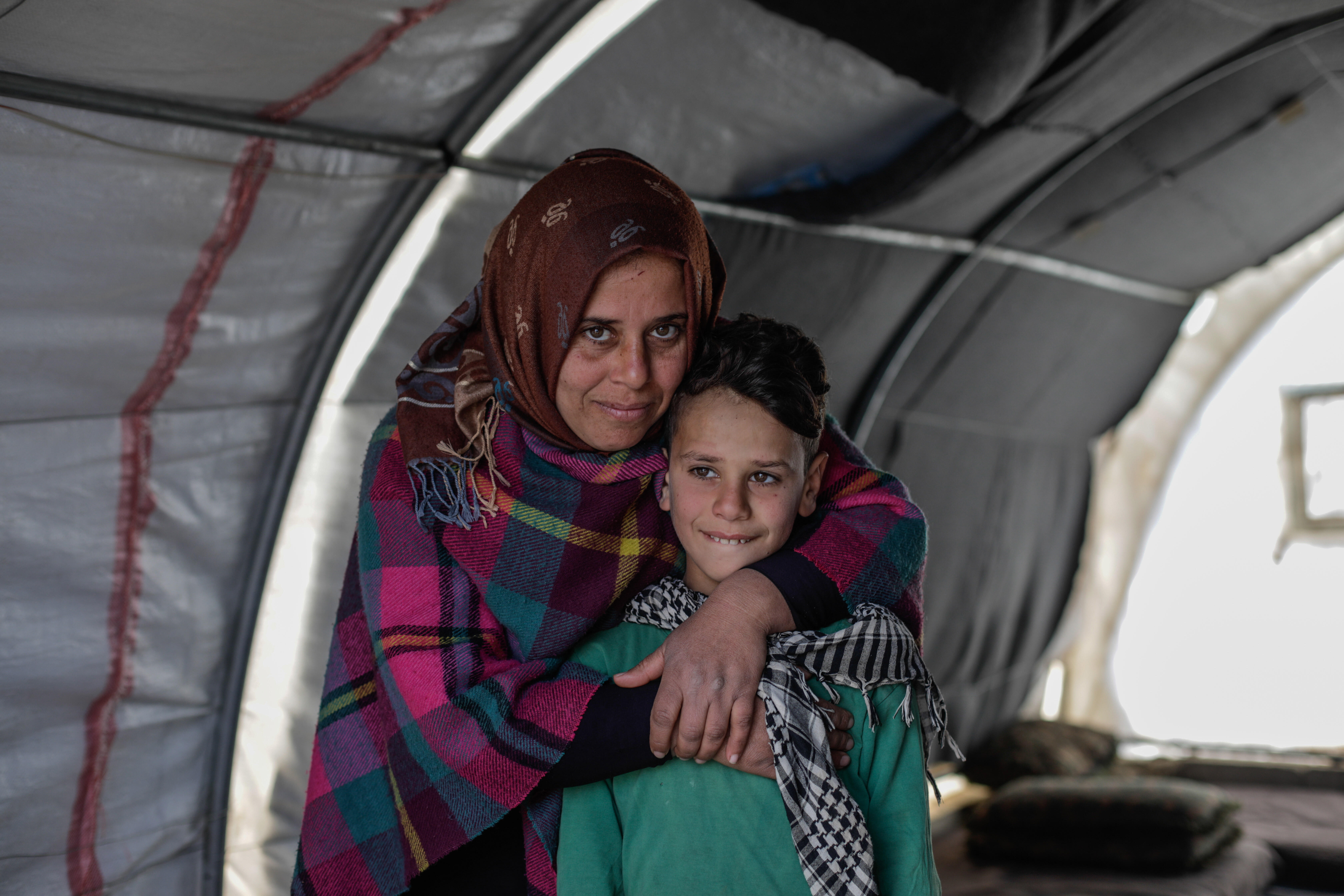 Sundus hugs her son Omar while standing behind him in a tent. Both are smiling and looking at the camera.