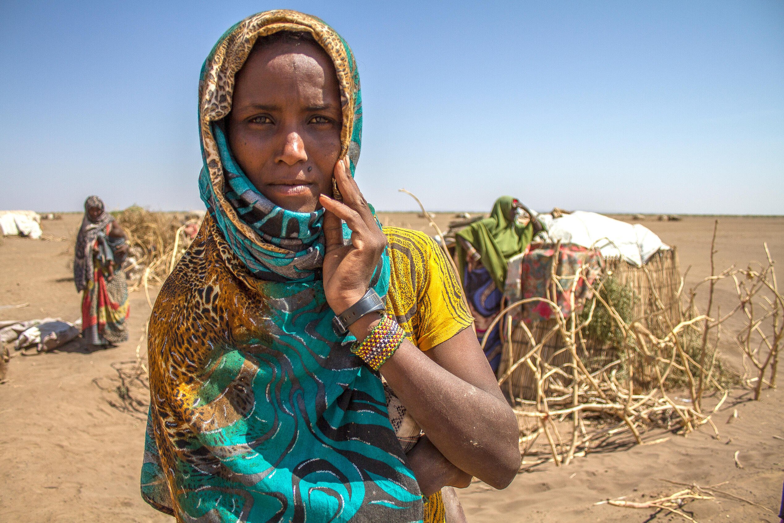 Deqa Omer, 32, stands among makeshift tents in a parched landscape in Ethiopia during a drought, her fingers resting on her cheek as she looks at the camera.