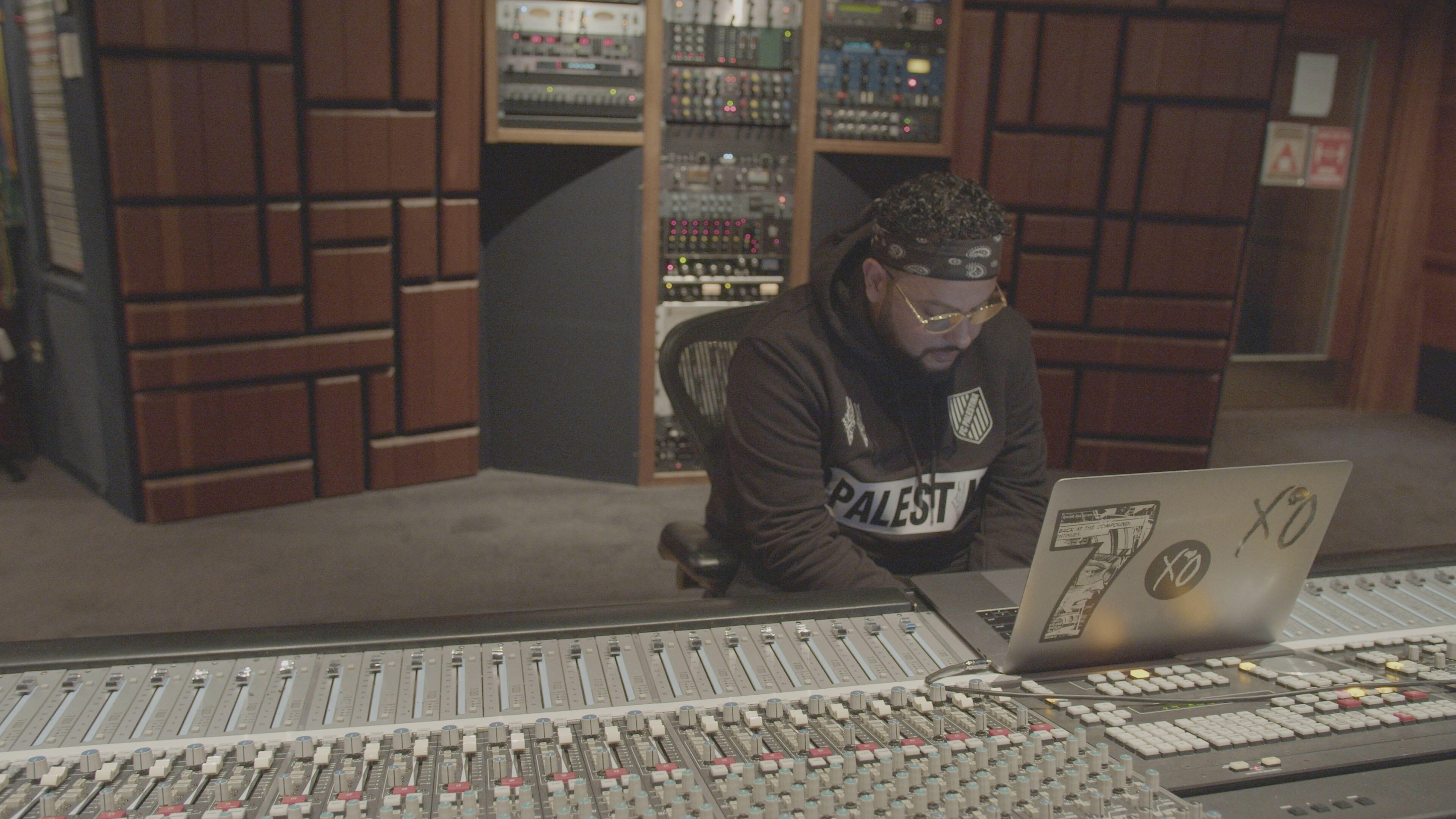 Belly, wearing a headband and a "Palestine" sweatshirt,  sits at his laptop, which is perched on a mixing board in a recording studio. 