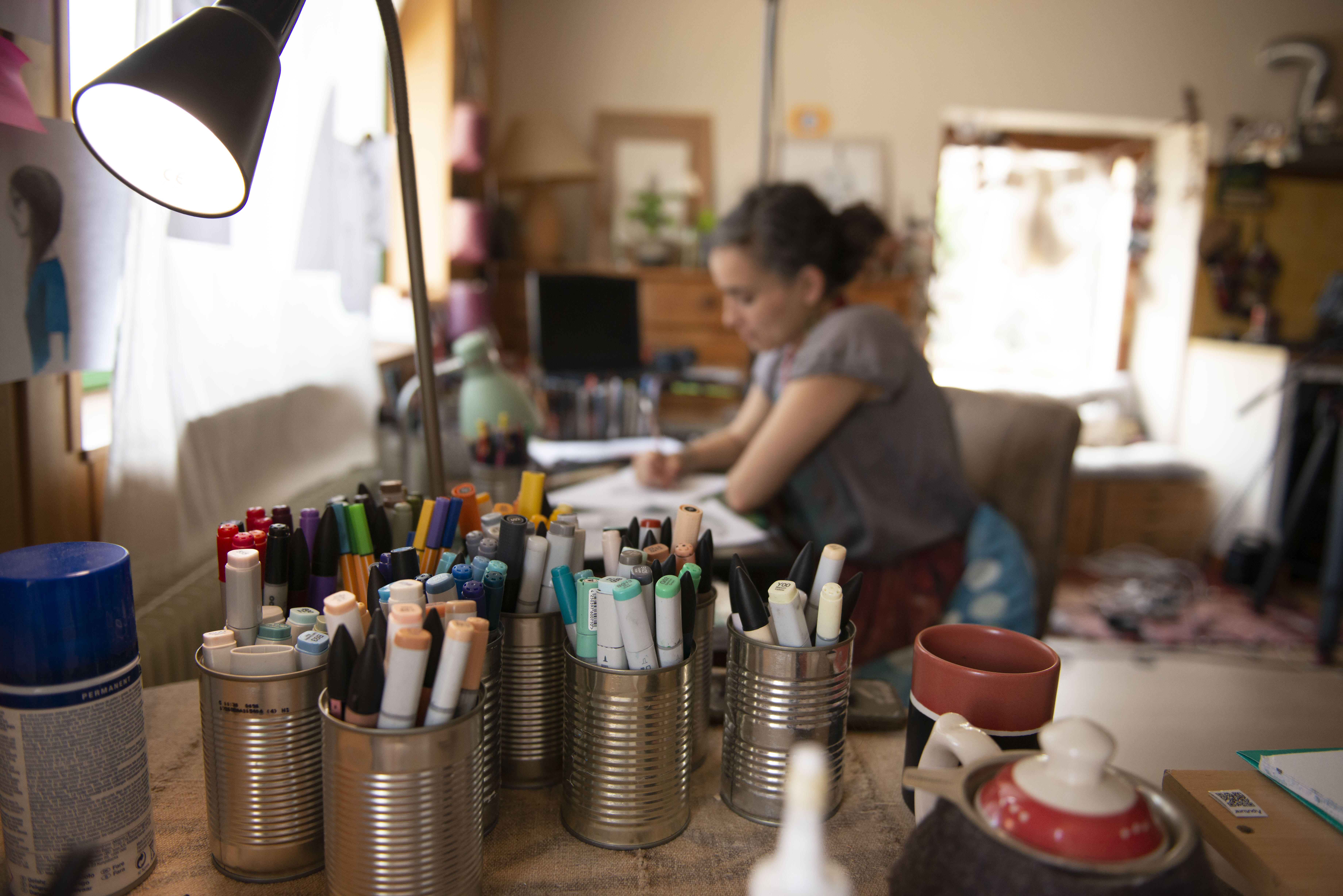 Diala Brisly sits at her drafting table, sketching an illustration for World Refugee Day 2021. Tin cans filled with markers and pens are in the foreground. 