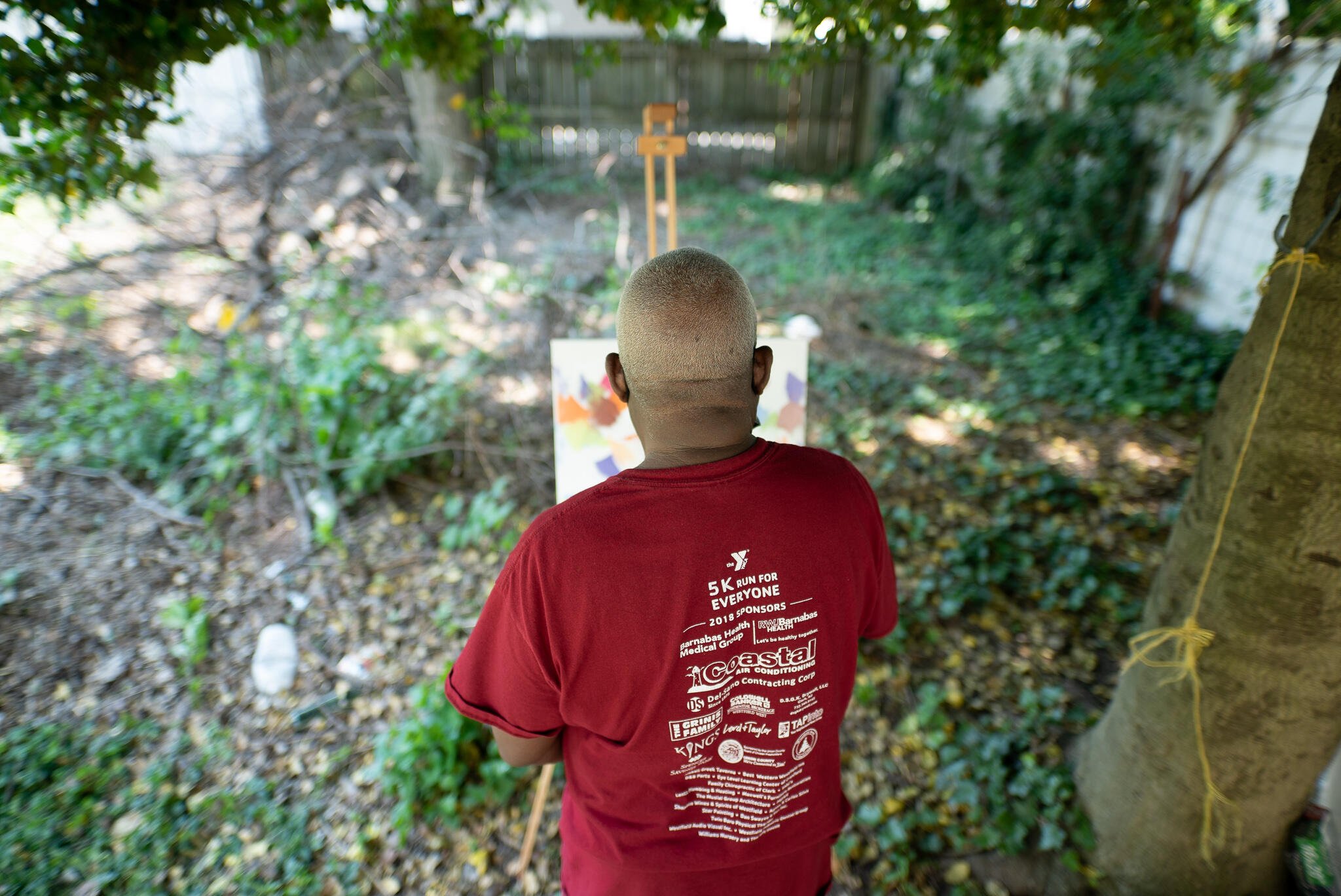 Congolese artist Muyambo Marcel Chishimba, seen from the back wearing a t-shirt, paints an oil painting at an easel in his New Jersey backyard