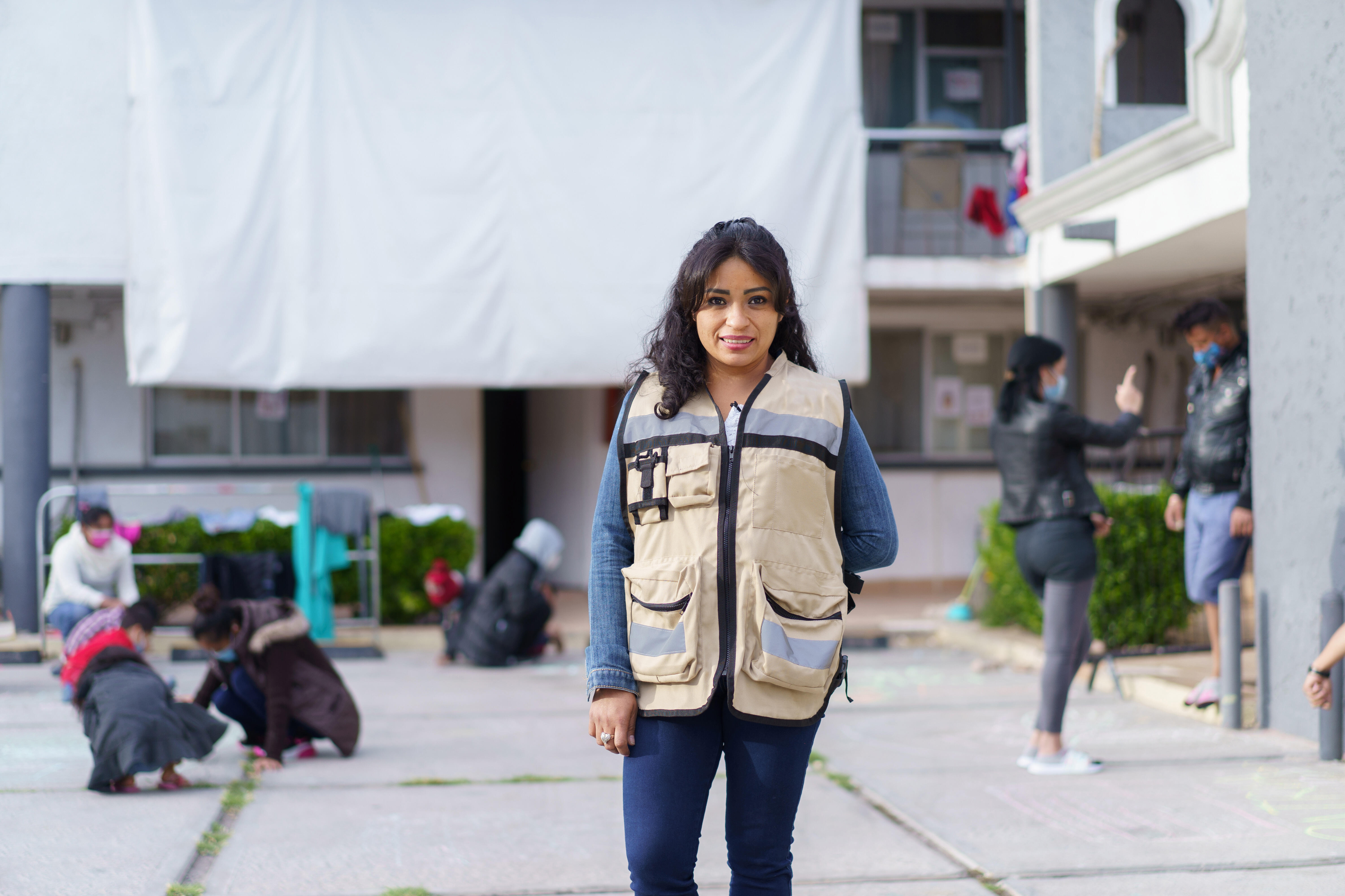 Rosa, wearing a brown vest with large pockets, stands in front of the triage hotel she helps manage in Ciudad Juárez, Mexico. In the background, children draw with chalk on the ground with their parents while other adults converse. 