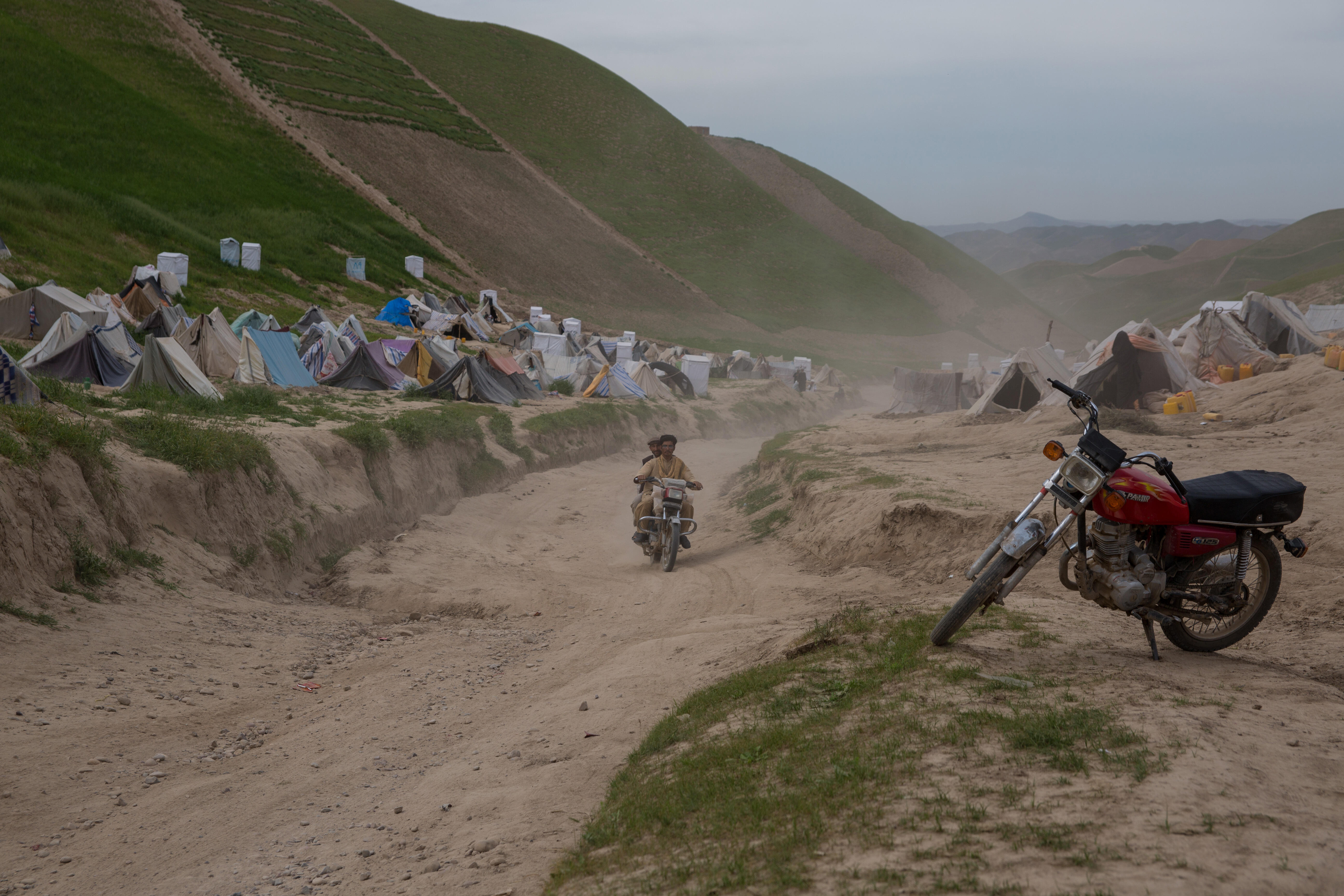 A camp for drought displaced people in Afghanistan. there is a motorcycle in the foreground as well as two men on a motorcycle on the road, and tents in the background on a mountainous landscape.