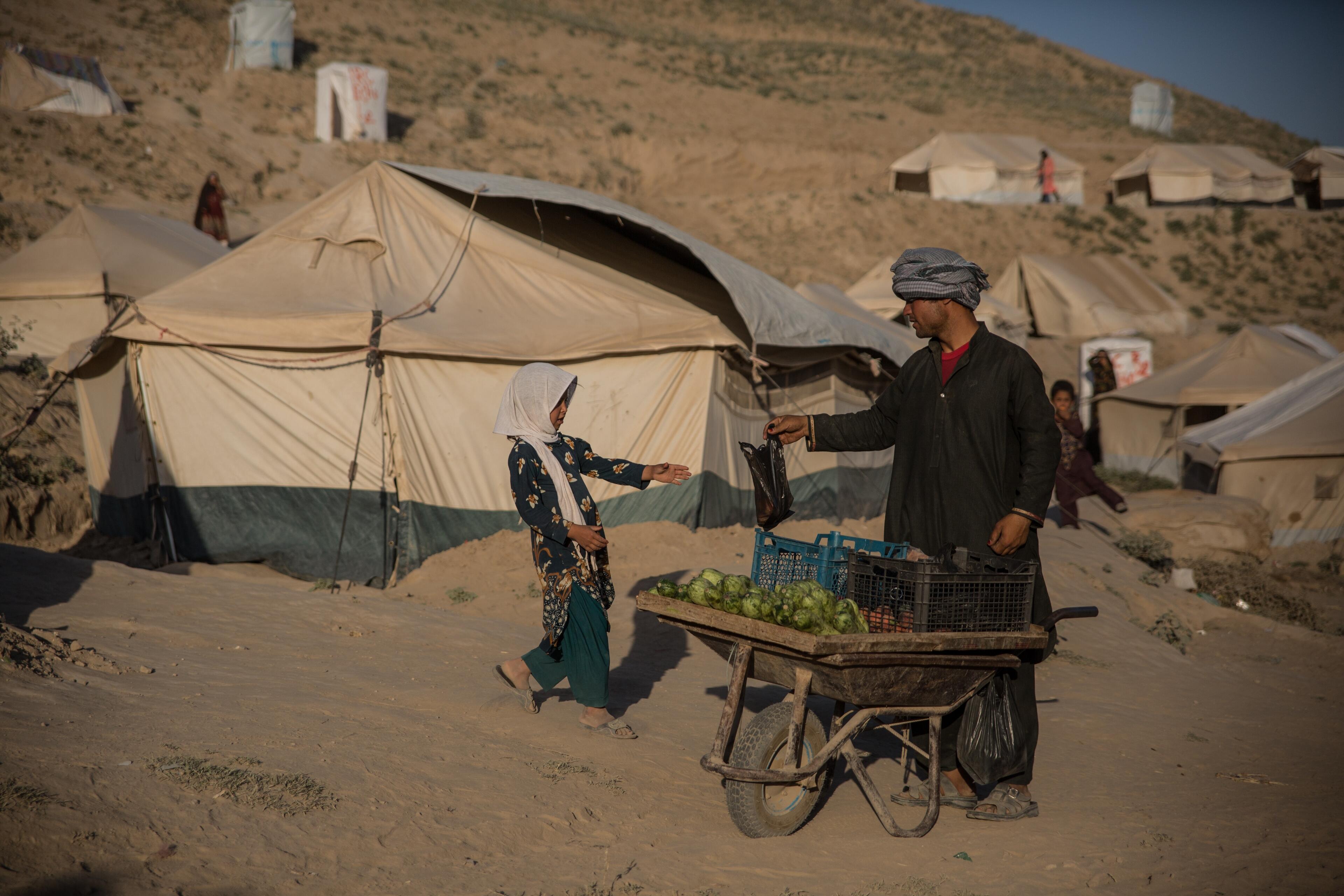 A man pushing a wheelbarrow with vegetables through a tent camp for displaced families in Badghis, Afghanistan hands a girl a plastic bag.
