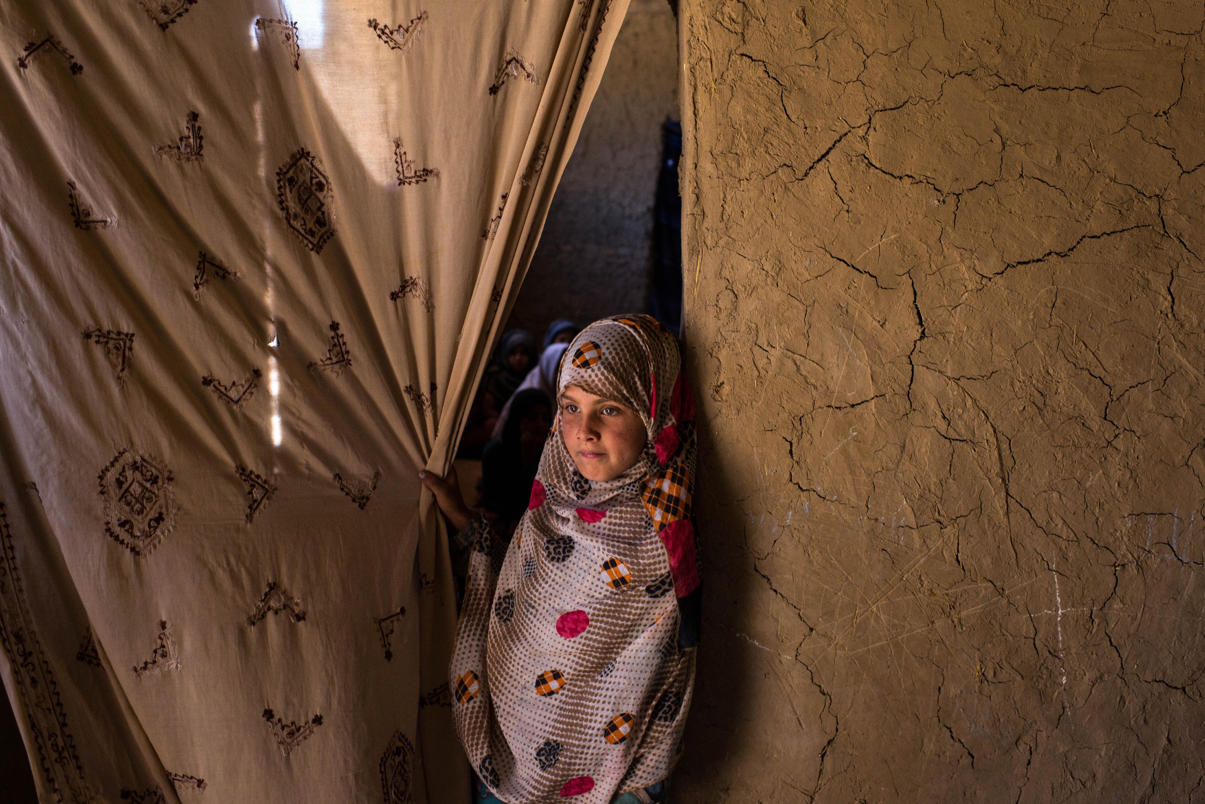 A young girl grasps a curtain hanging in a doorway in Afghanistan, looking out as she leans agains the wall.