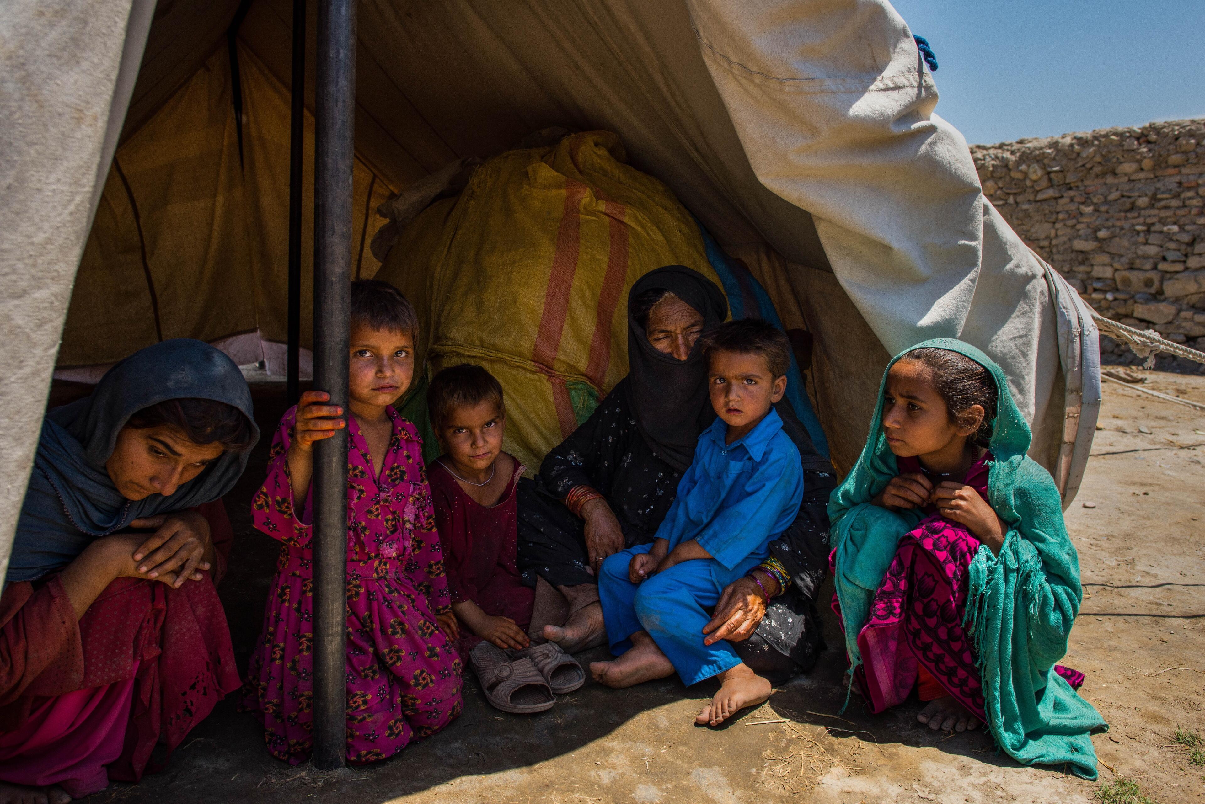 Women and children from an Afghan family living in a displacement camp sit inside the shade of their tent in an arid landscape.