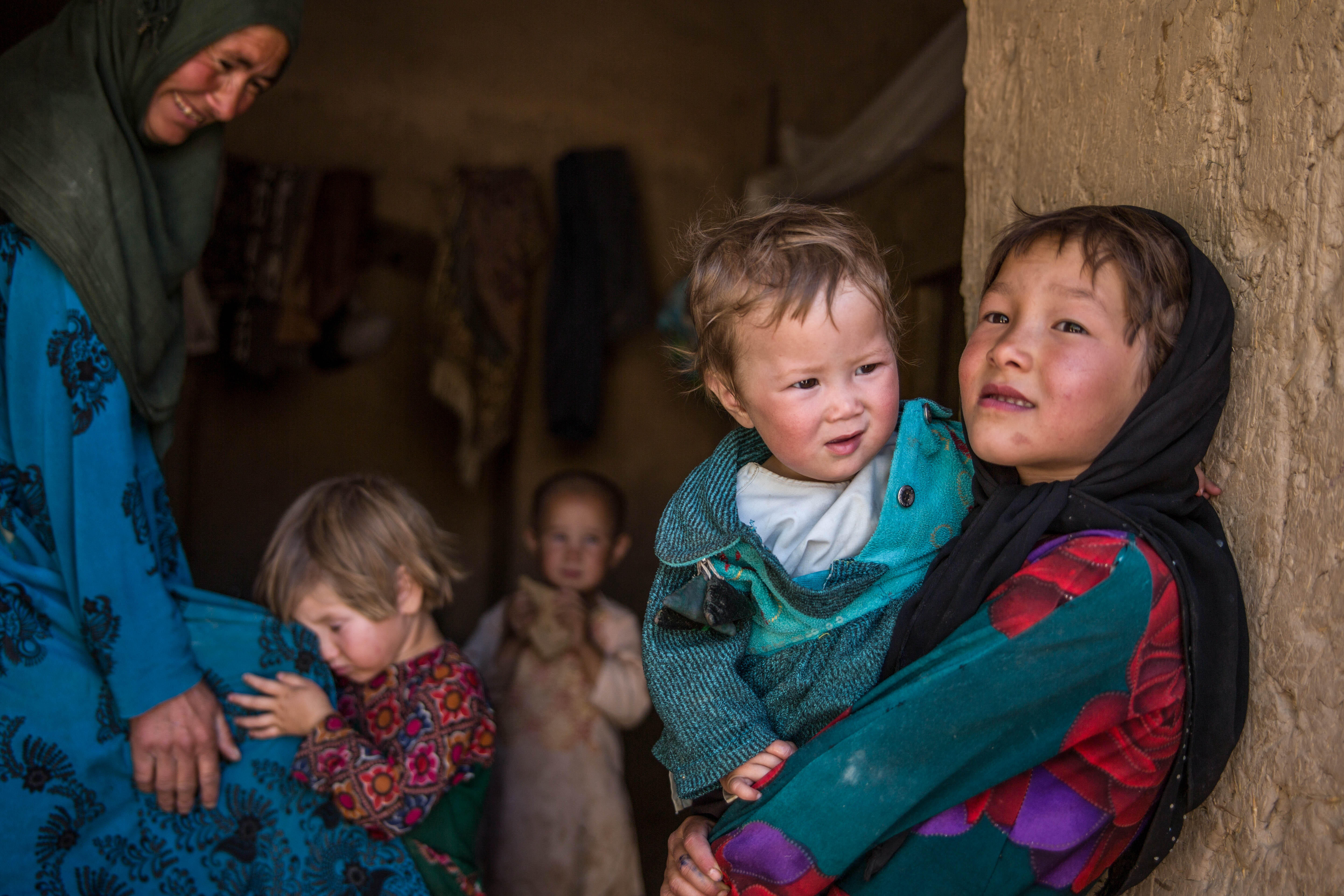 A young Afghan girl holds a younger sibling. Her mother and two other children are in the background. 