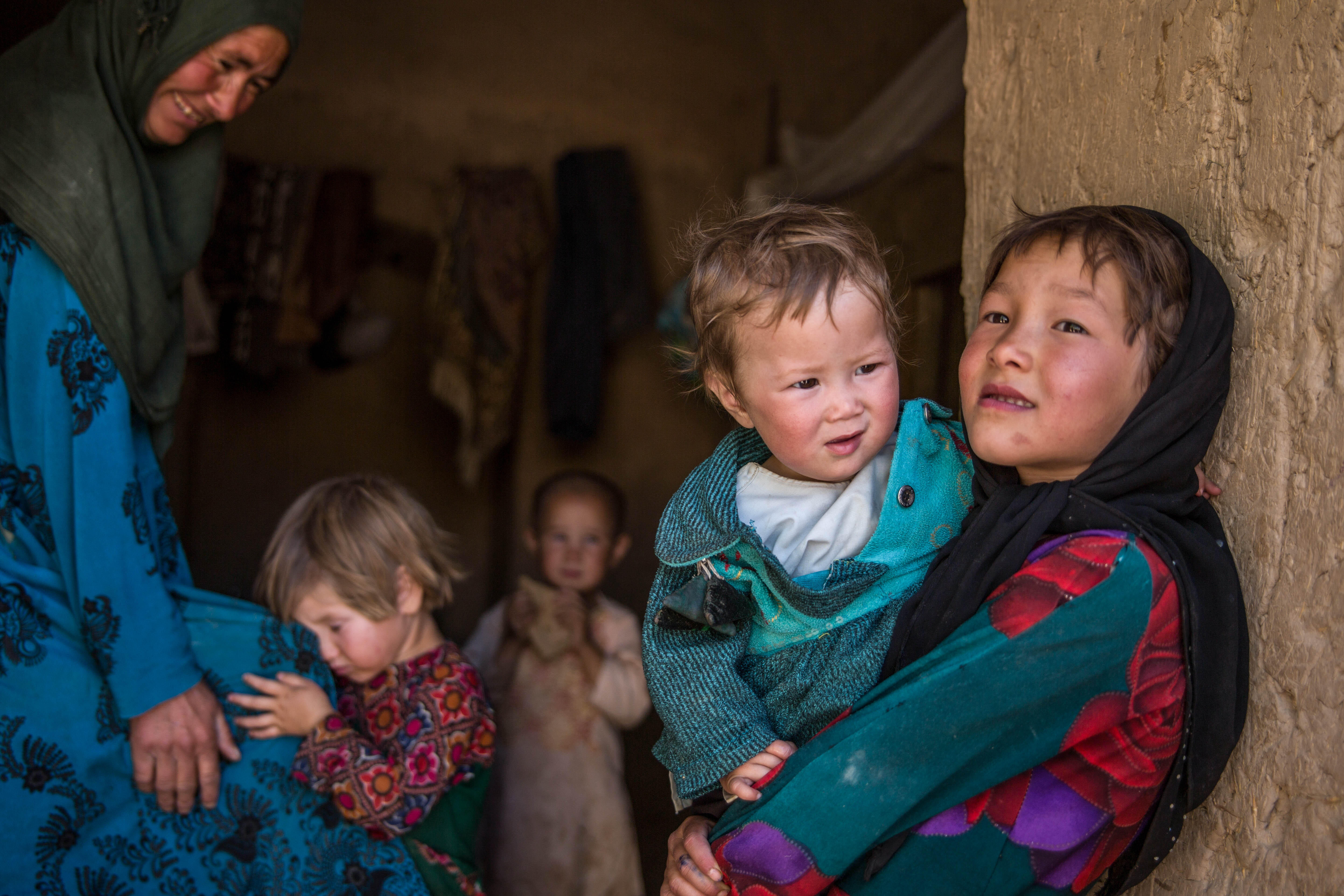 A young Afghan girl holds a younger sibling. Her mother and two other children are in the background.