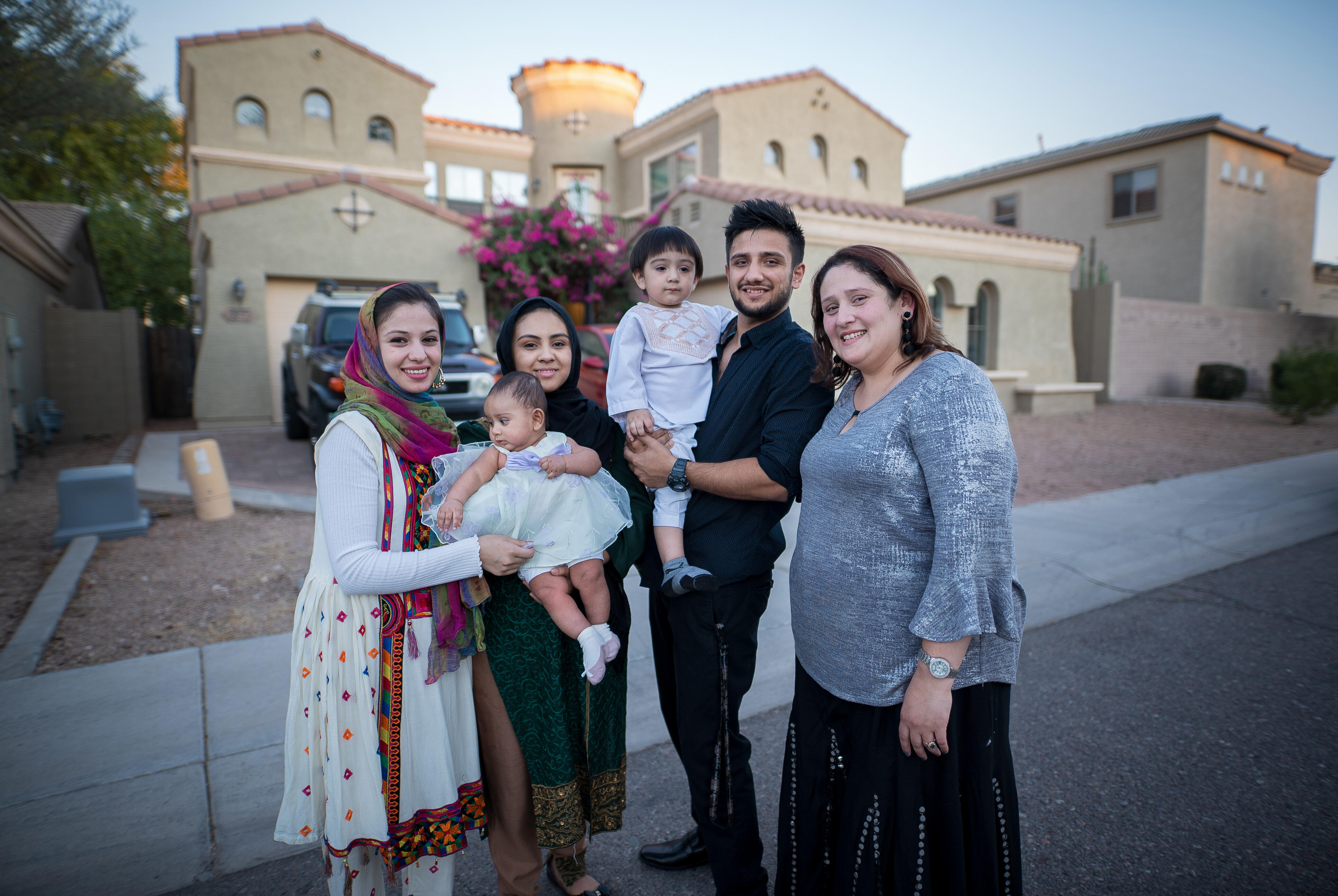 Muska, a woman in her twenties, stands in front of her home with three adult family members, a toddler and a baby. 