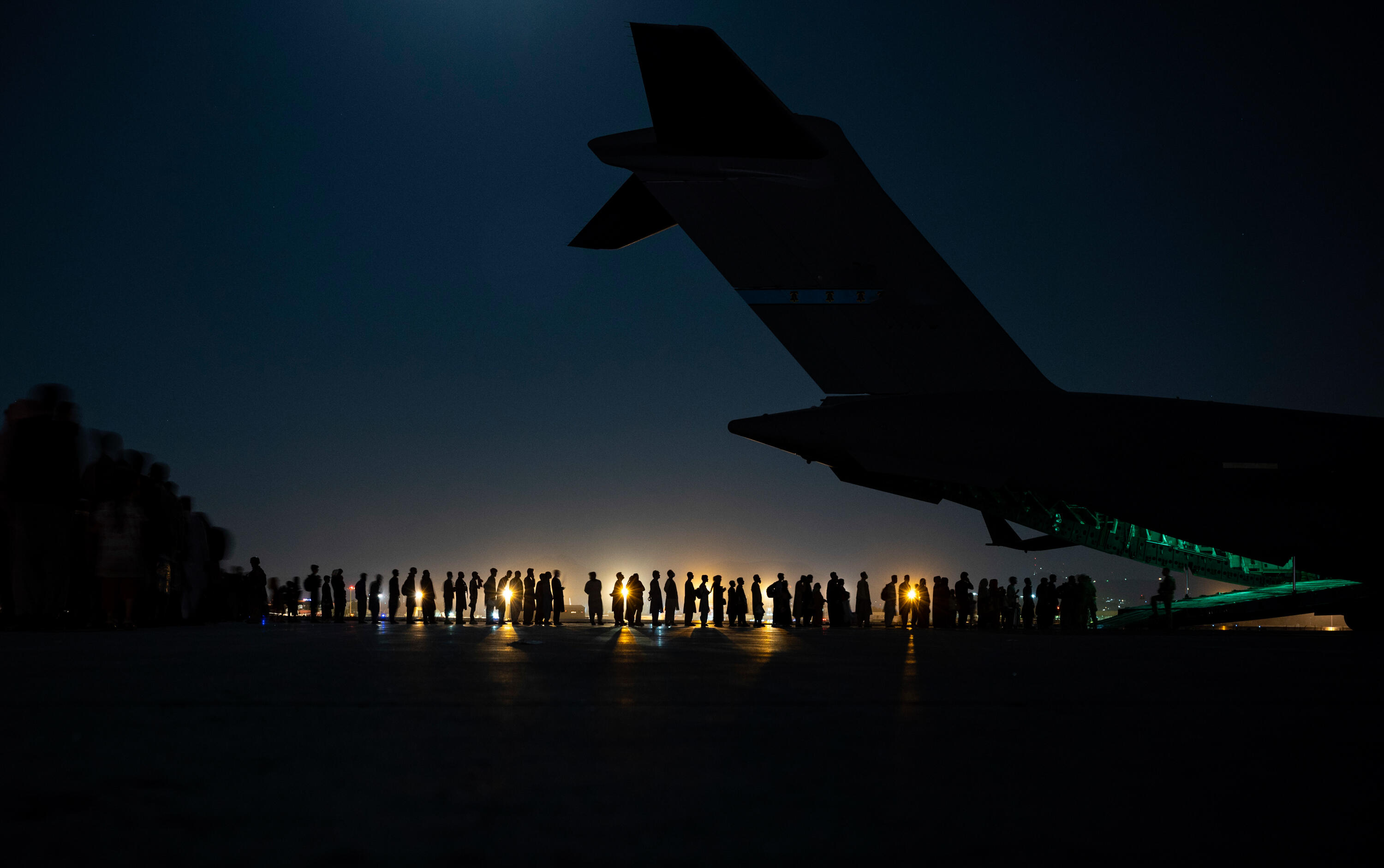 A night shot, with the tail of an airplane in the foreground and a line of people in the background. We can see a light in the distance and just the silhouette of the plane and people.