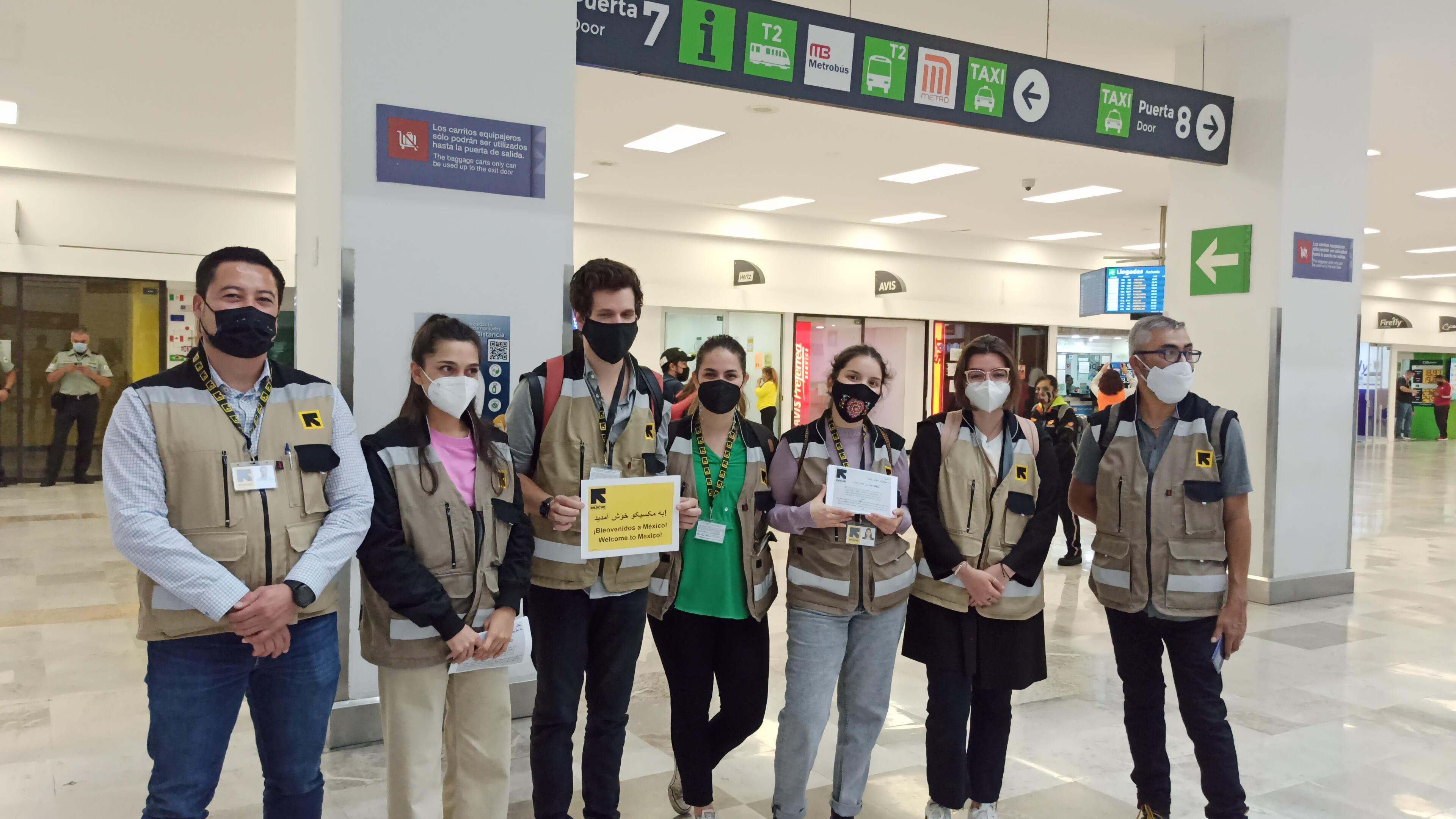 IRC staff members, all wearing masks and IRC-branded vests, stand in a line in an airport terminal. Some are holding welcome signs.
