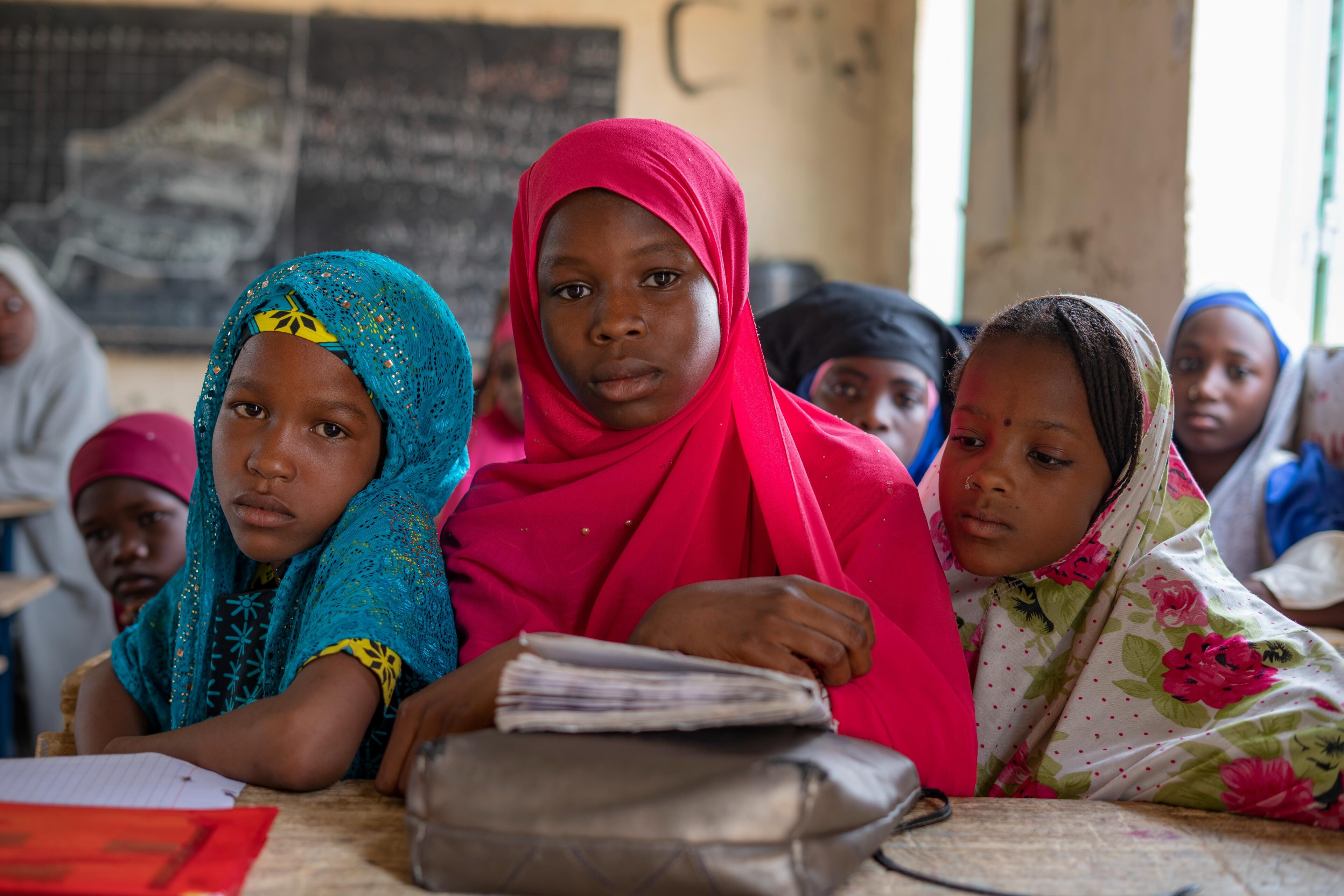 Fifteen-year-old Maryama sits with a serious expression at a school desk with two friends. A school book and papers are on the desk in front of the girls.