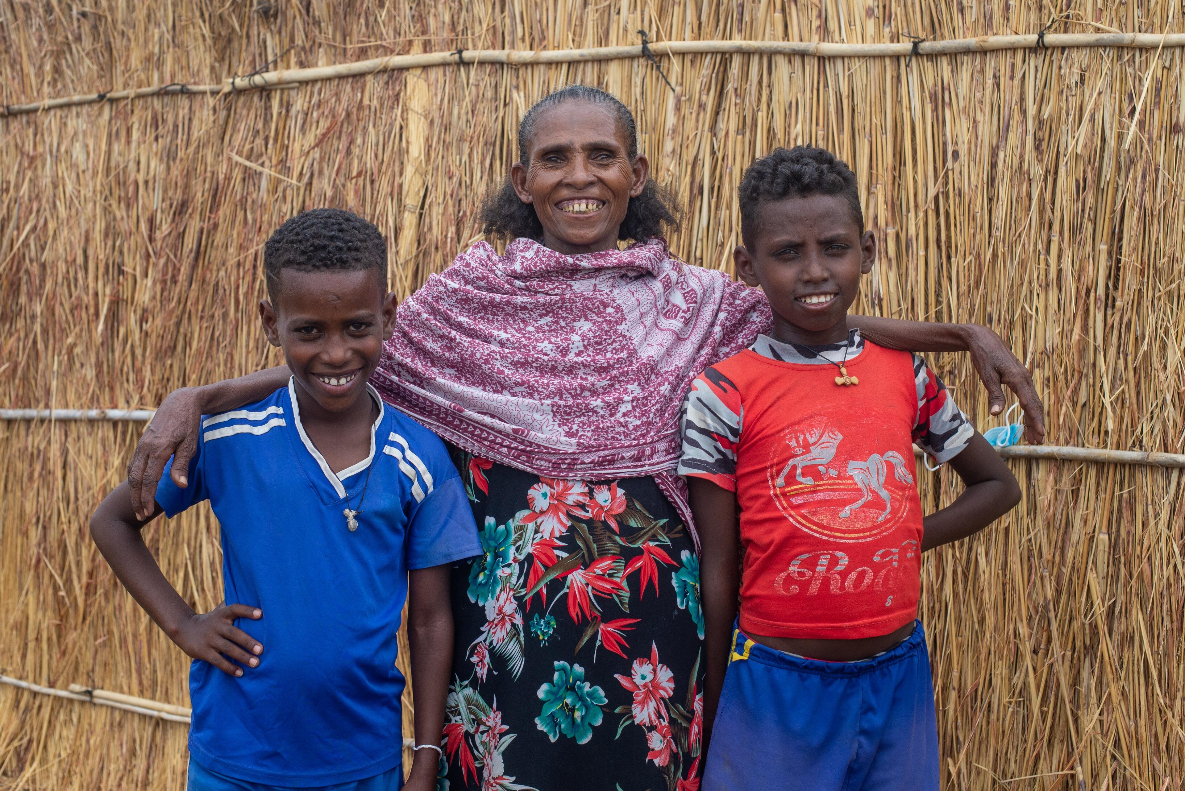 A Tigrayan boy named Hafton stands smiling with his mother Wahid and his best friend Mherka outside a grass-walled shelter in a refugee camp in eastern Sudan.