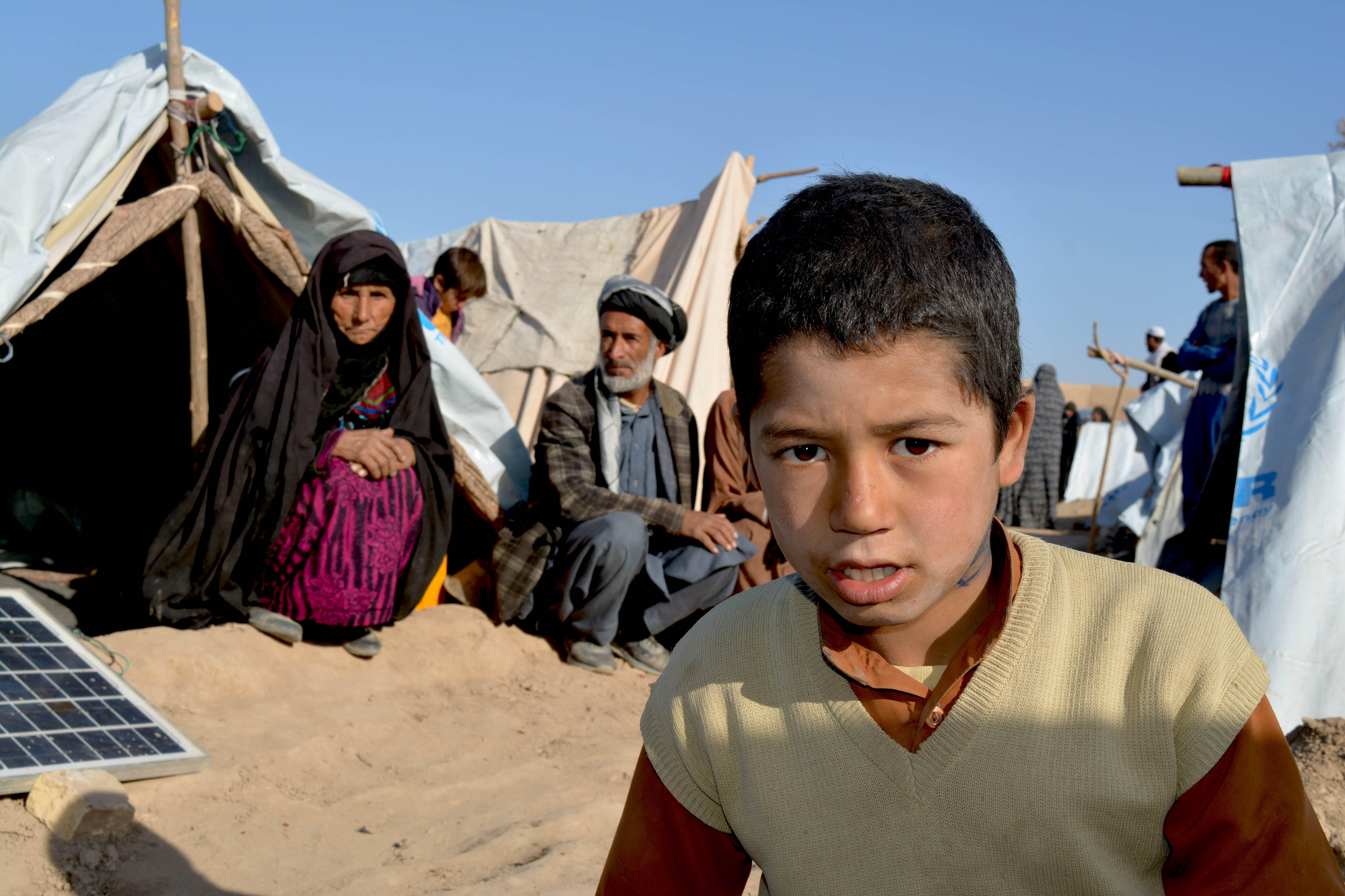 A boy leans toward the camera as an elderly man and woman sit on the sand in front of their tent in a camp for displaced families in Afghanistan.