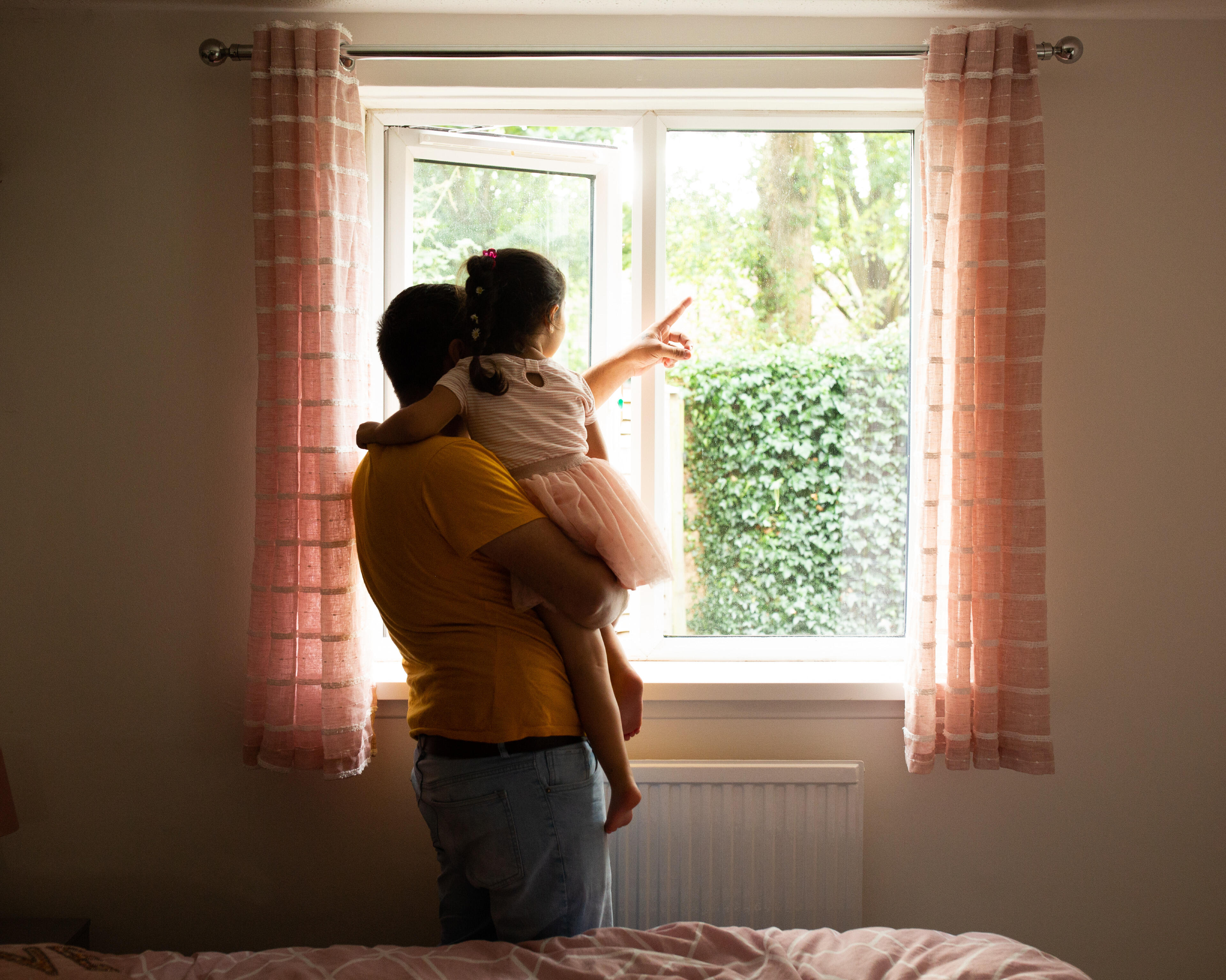 Maasom holds his 4-year-old daughter Nasrin, pointing at a tree as they look through a window at a garden.