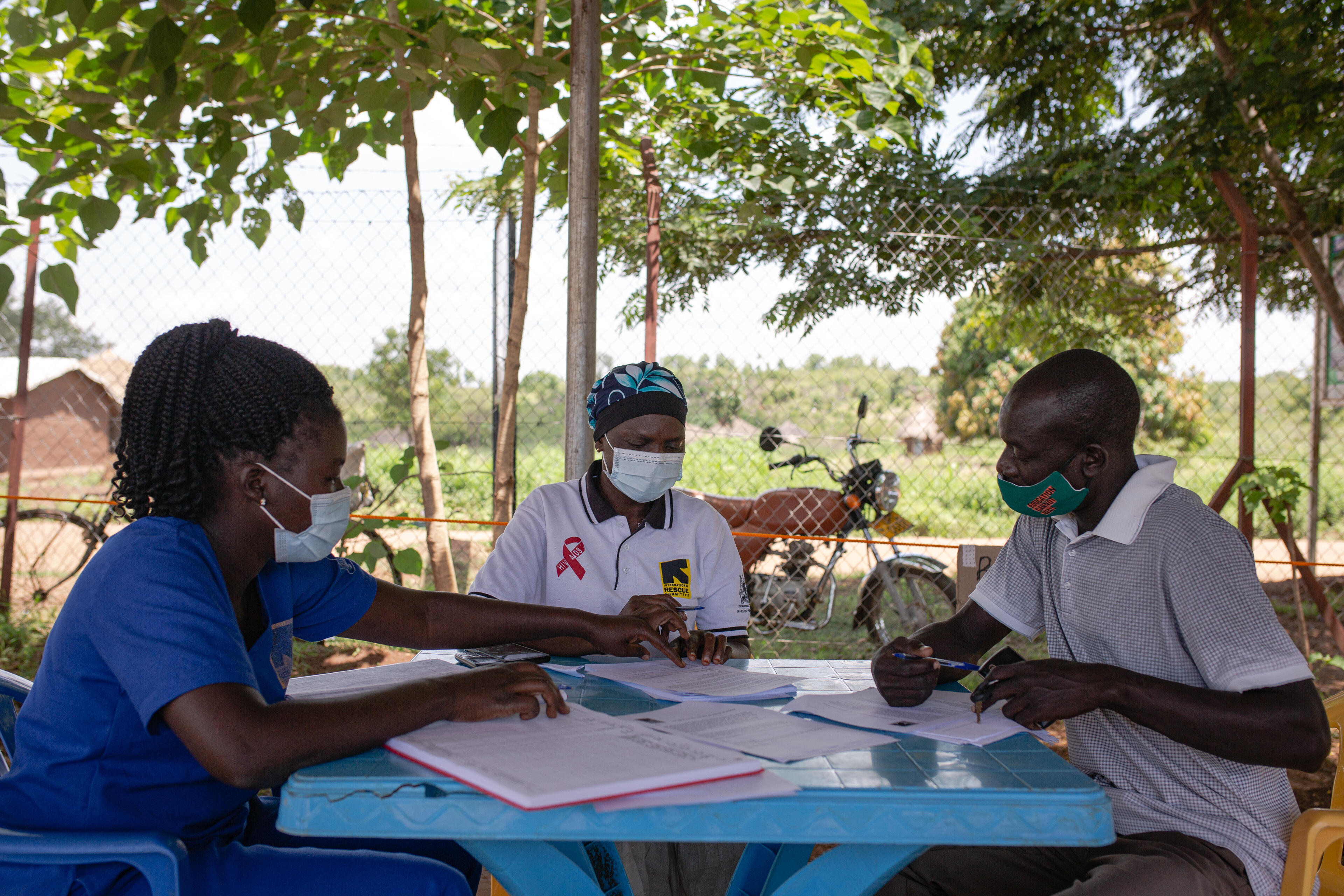 Ugandan teacher Azilu Rasulu sits at a table outdoors listening to two female IRC staff provide information about the COVID-19 vaccine.