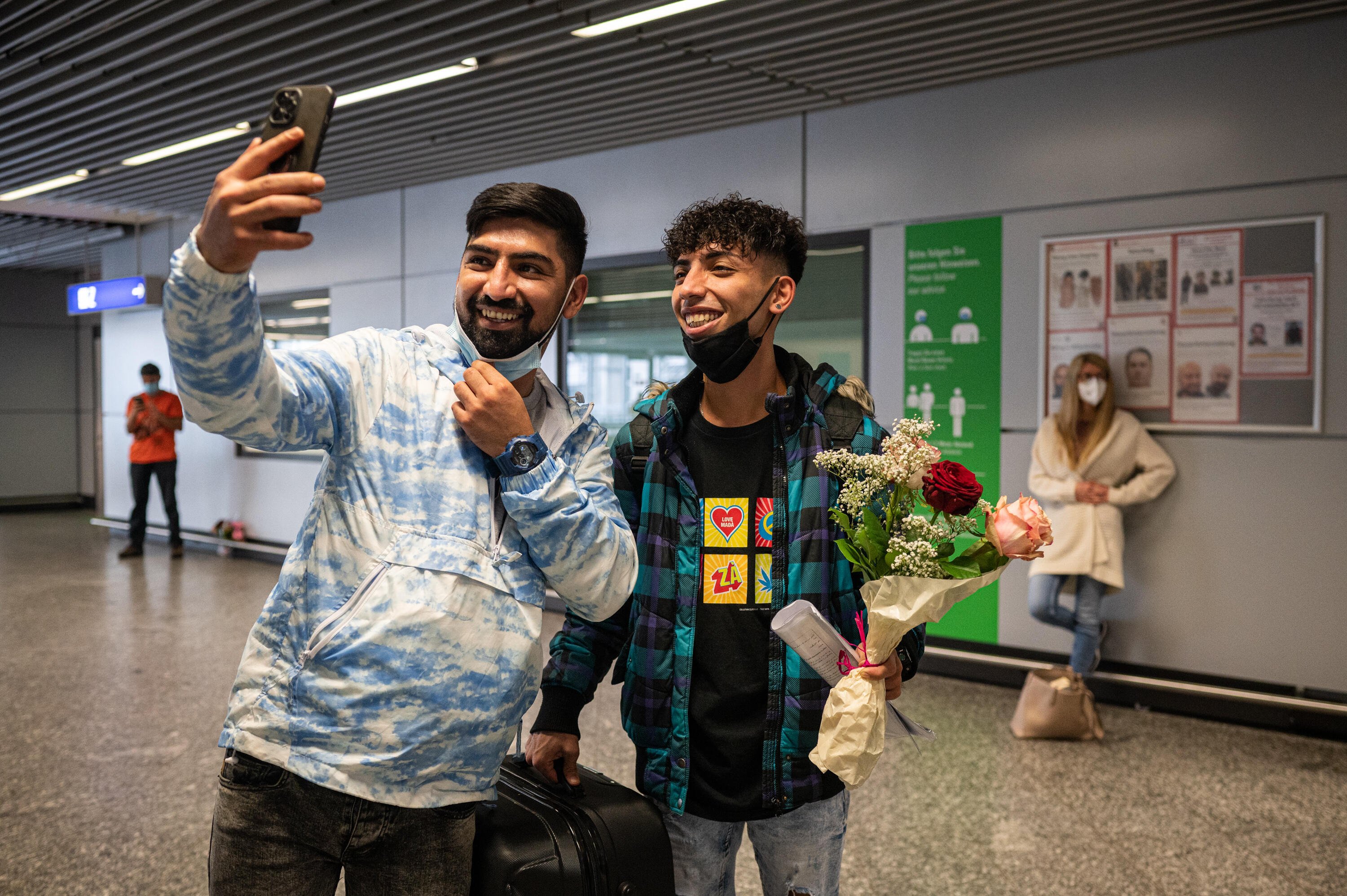 In an airport, two young men look at a phone to take a selfie. They are smiling and one of them holds flowers. 