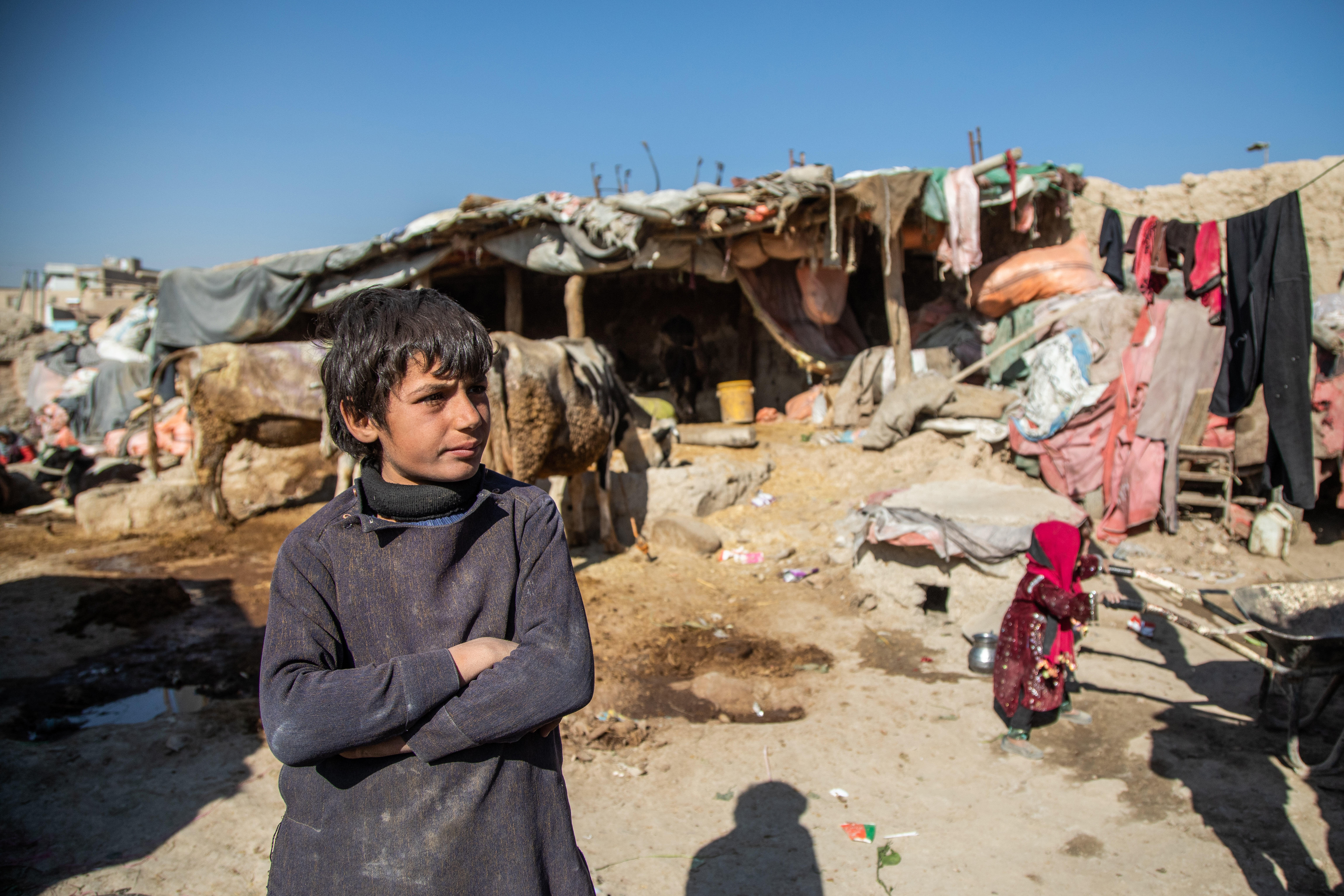 In front of a makeshift shelter, a boy stands with his arms crossed. 