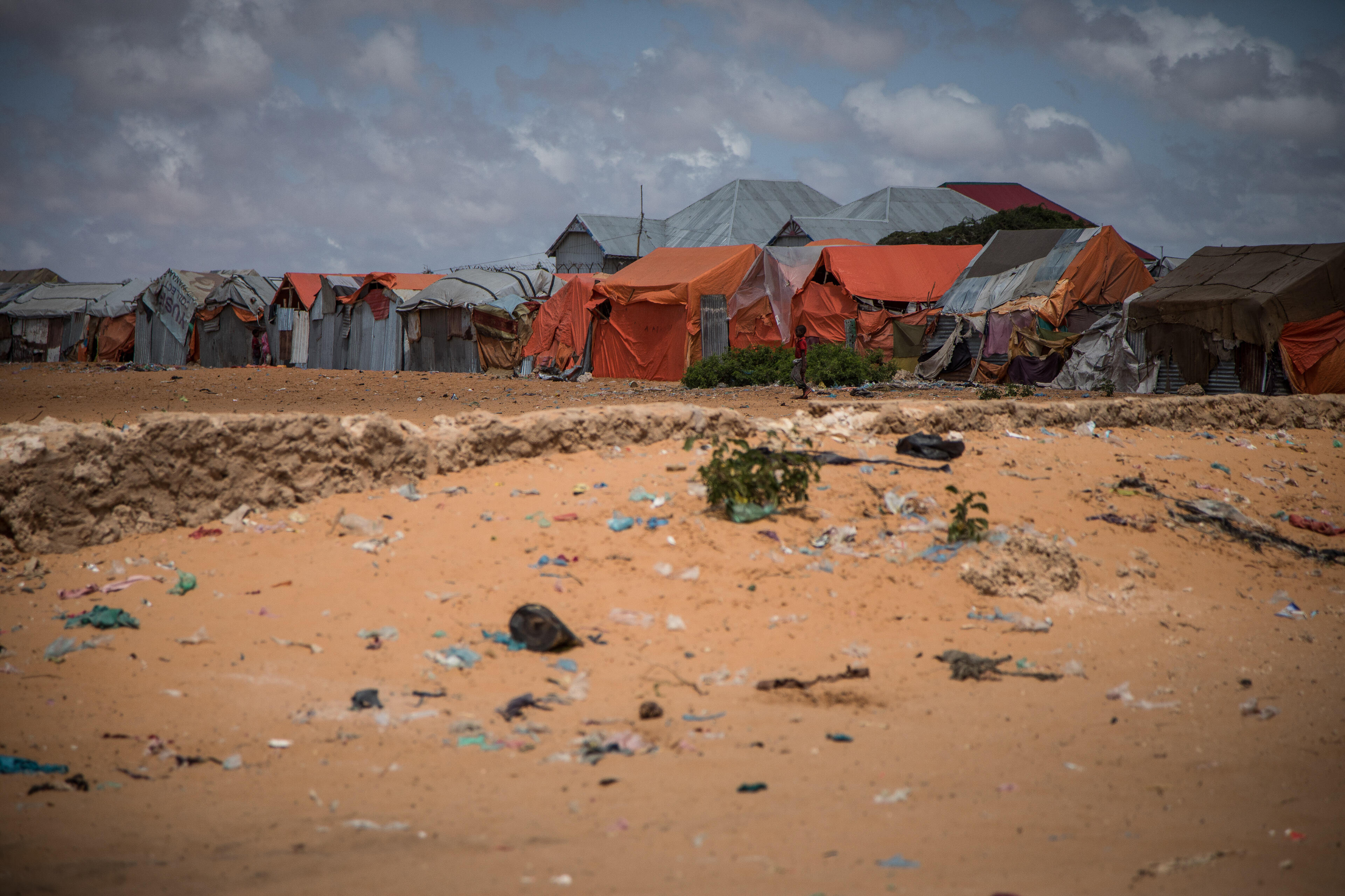 A small boy stands against the backdrop of tents for displaced families in an arid, littered landscape outside Mogadishu, Somalia.