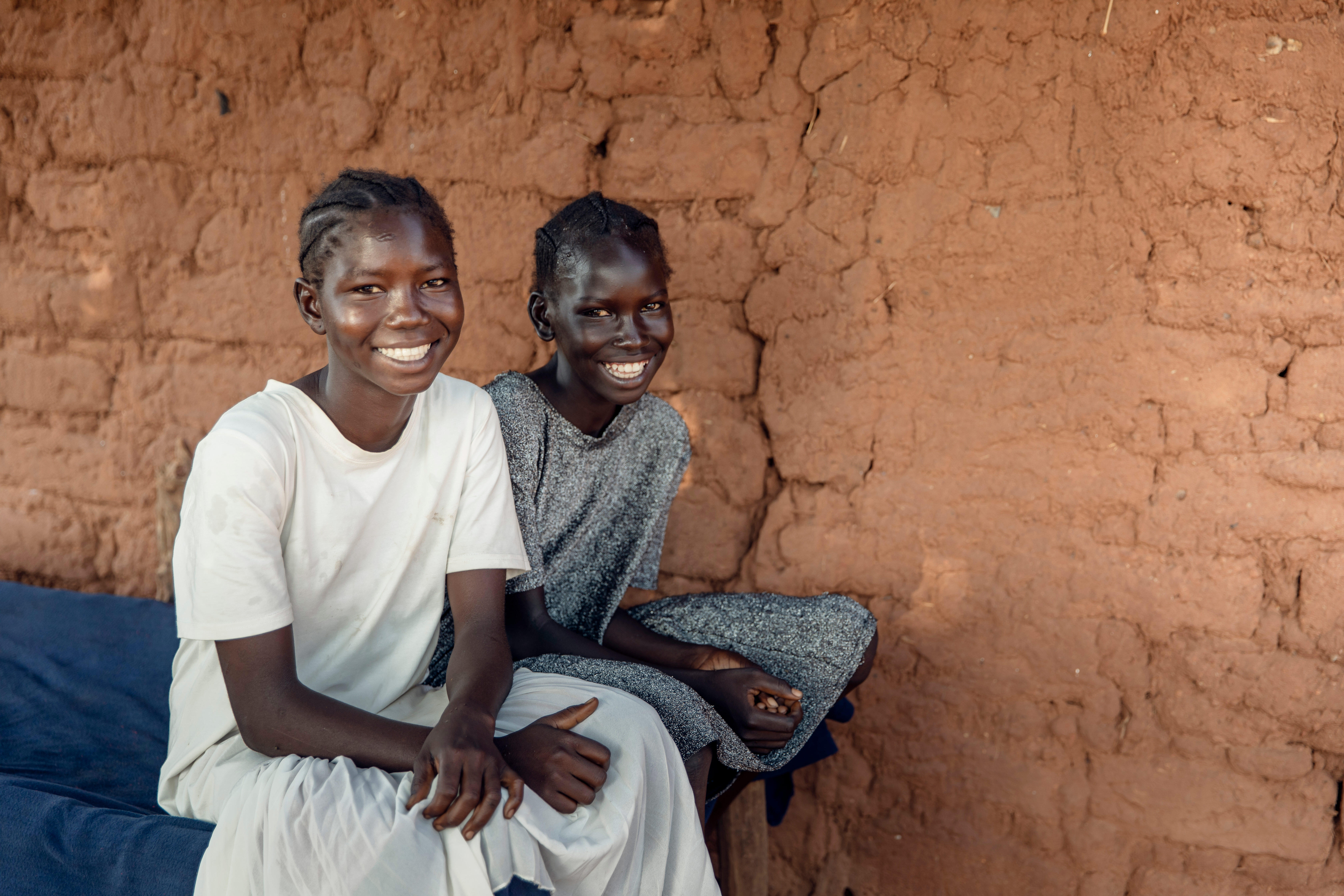 Two young girls sit on the edge of a bed and smile looking at the camera.