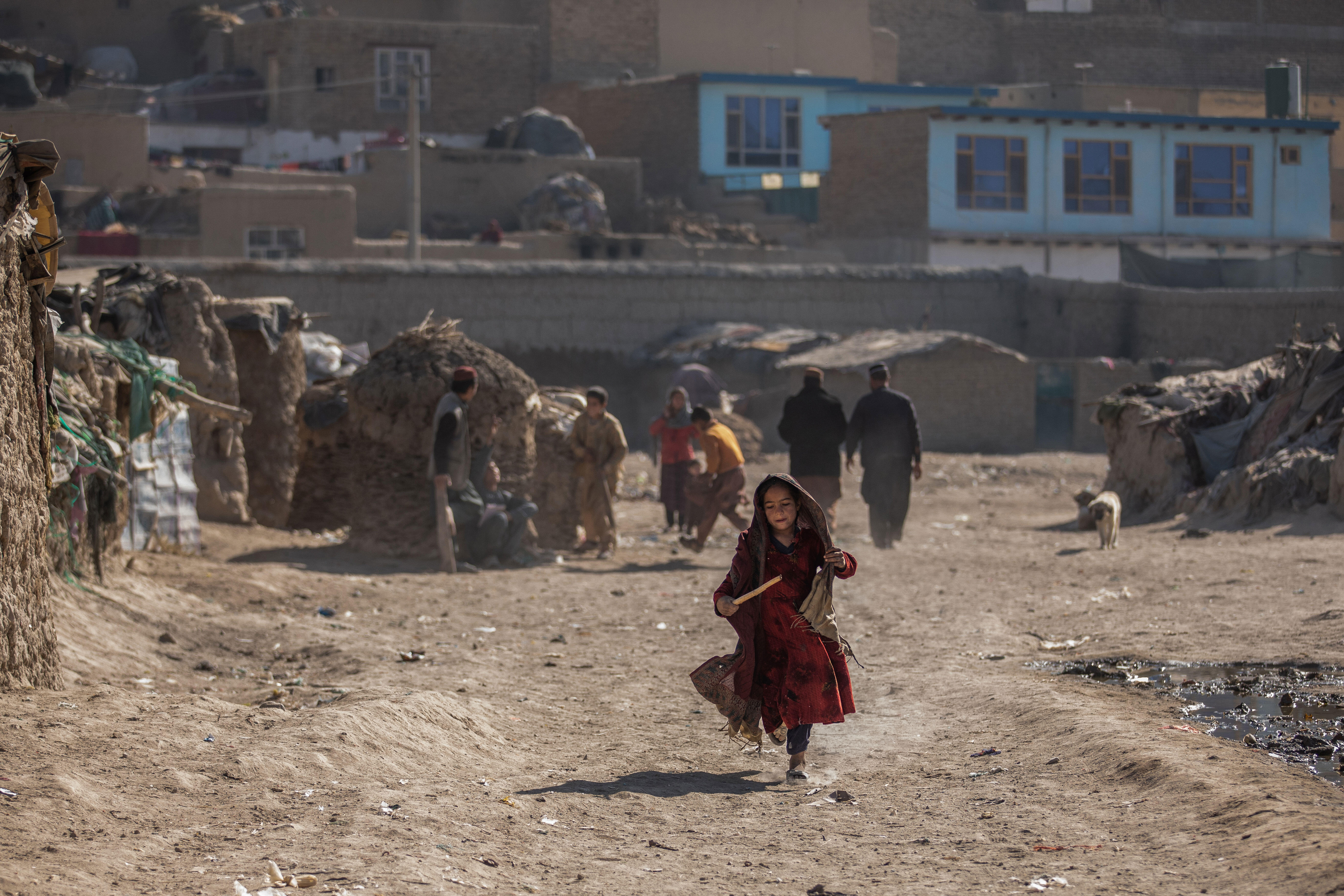 In a camp in Afghanistan, a young girl runs while holding a stick. There are other people and makeshift homes in the background.