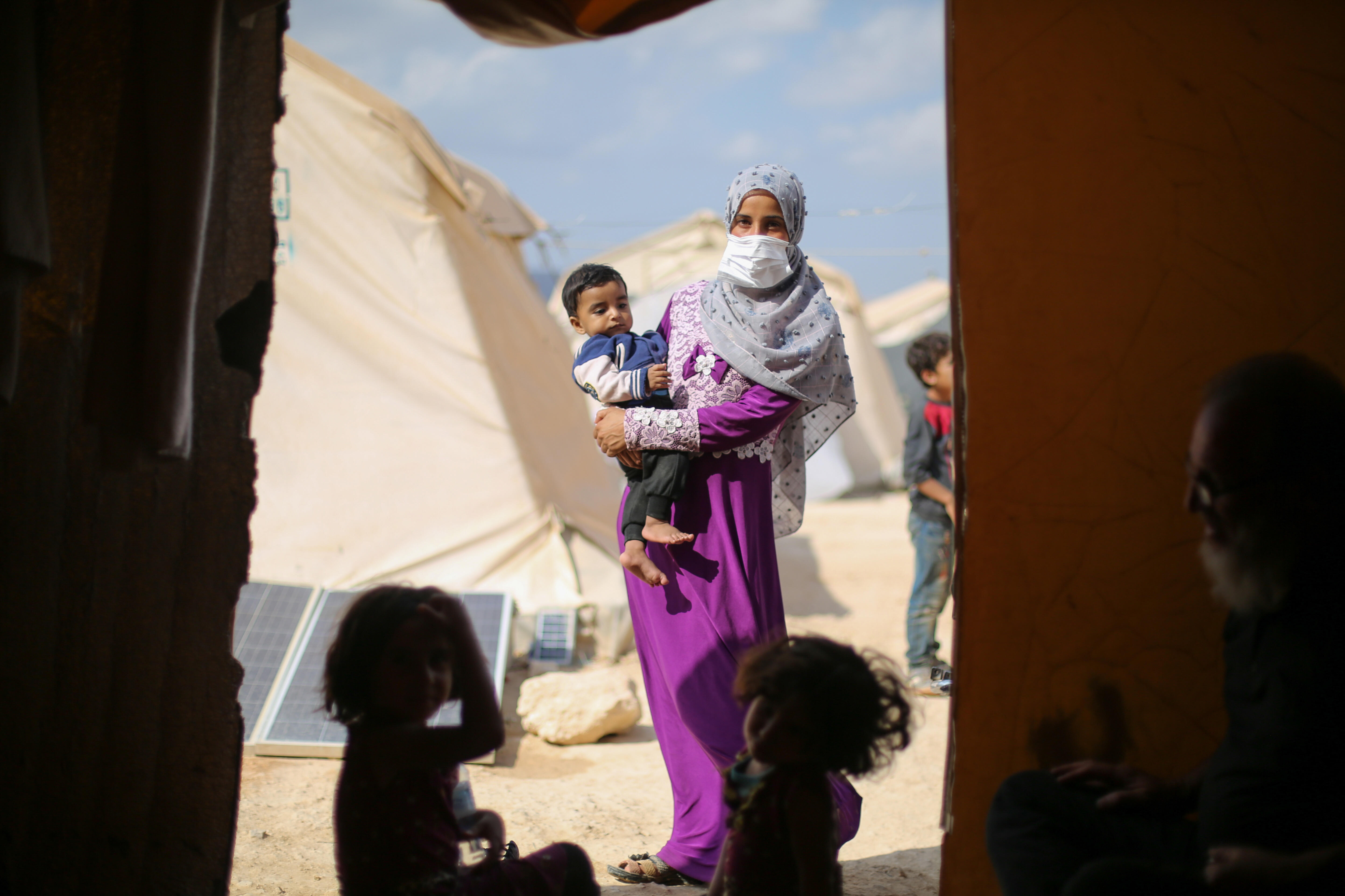 Two children sit on the ground in a tent. Framed in the entrance to the tent is a woman holding a toddler. There are other tents in the background.