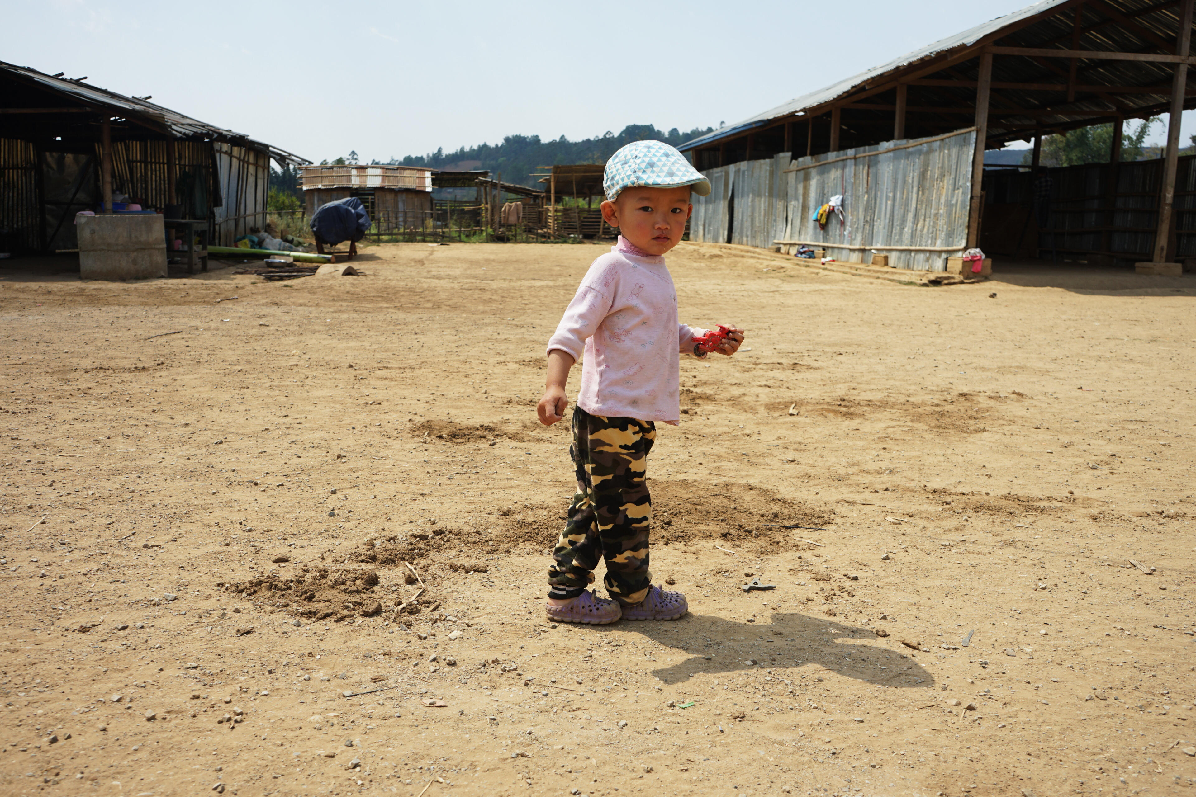 A toddler boy holds a small toy motorcycle as he stands in a wide dusty area between long, corrugated tin structures in a displacement camp.