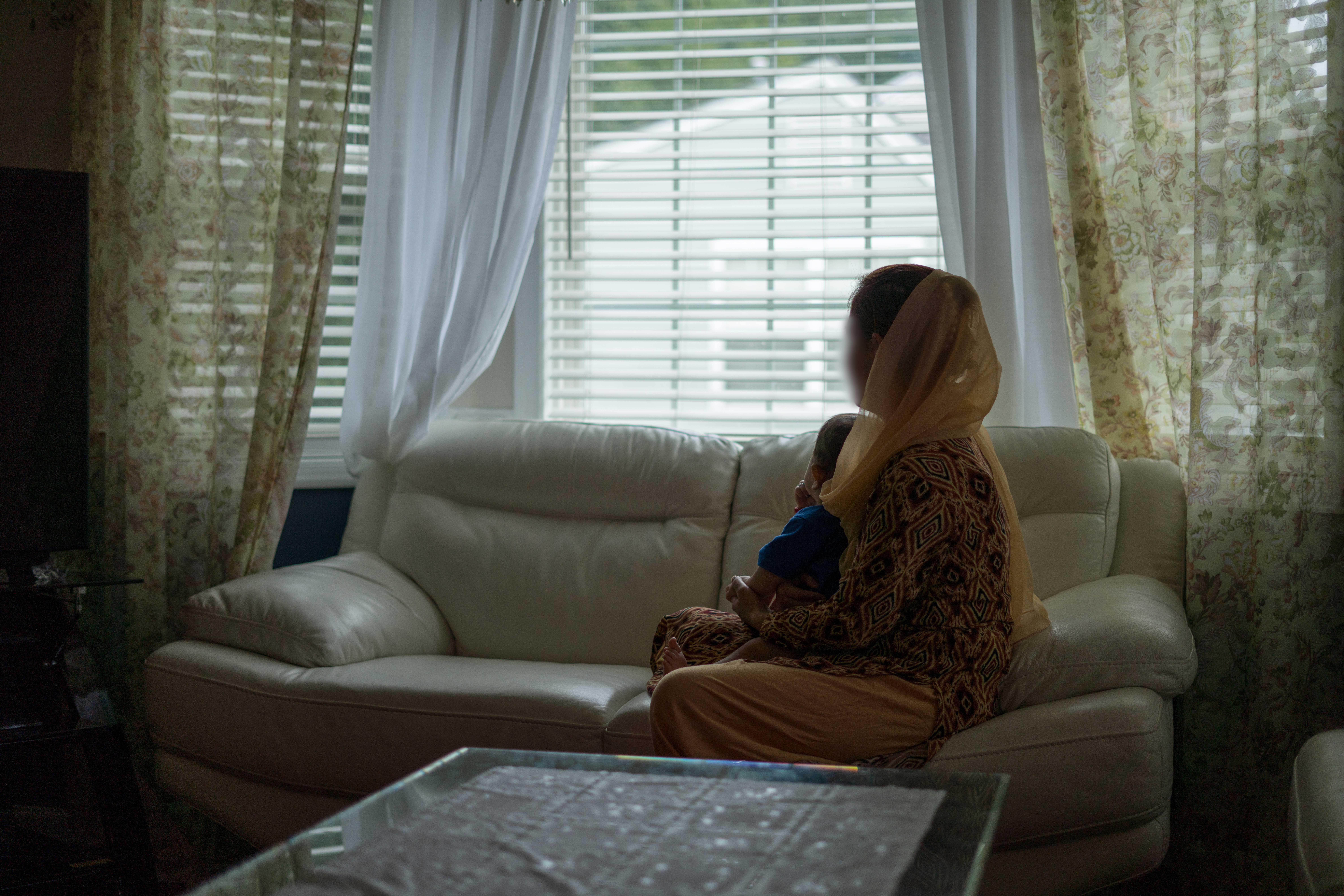 A woman sits on a couch in a living room looking out a window with a toddler on her lap. 