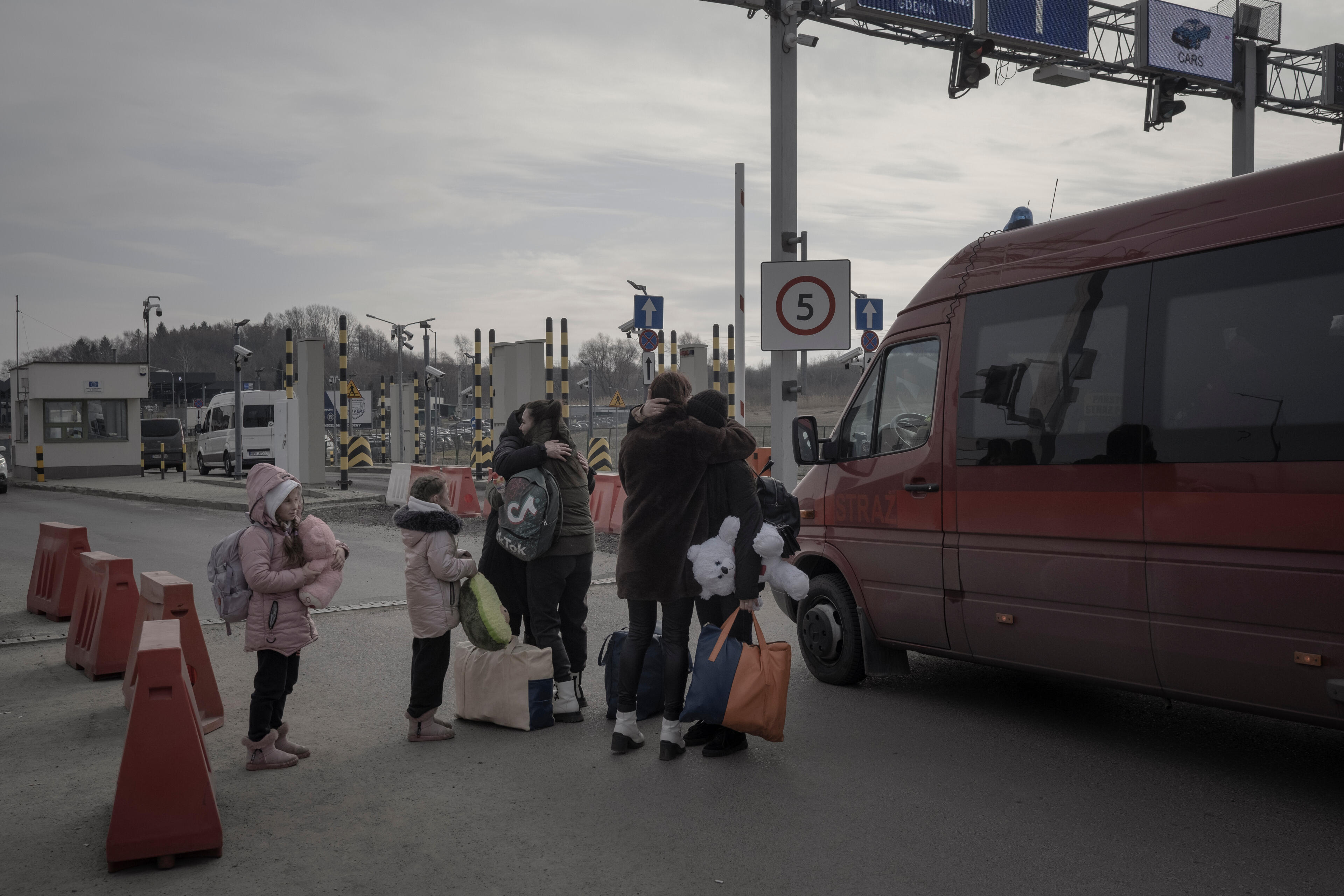 A Ukrainian family embraces at a Ukraine-Poland border crossing next to a van. One person holds a big teddy bear.