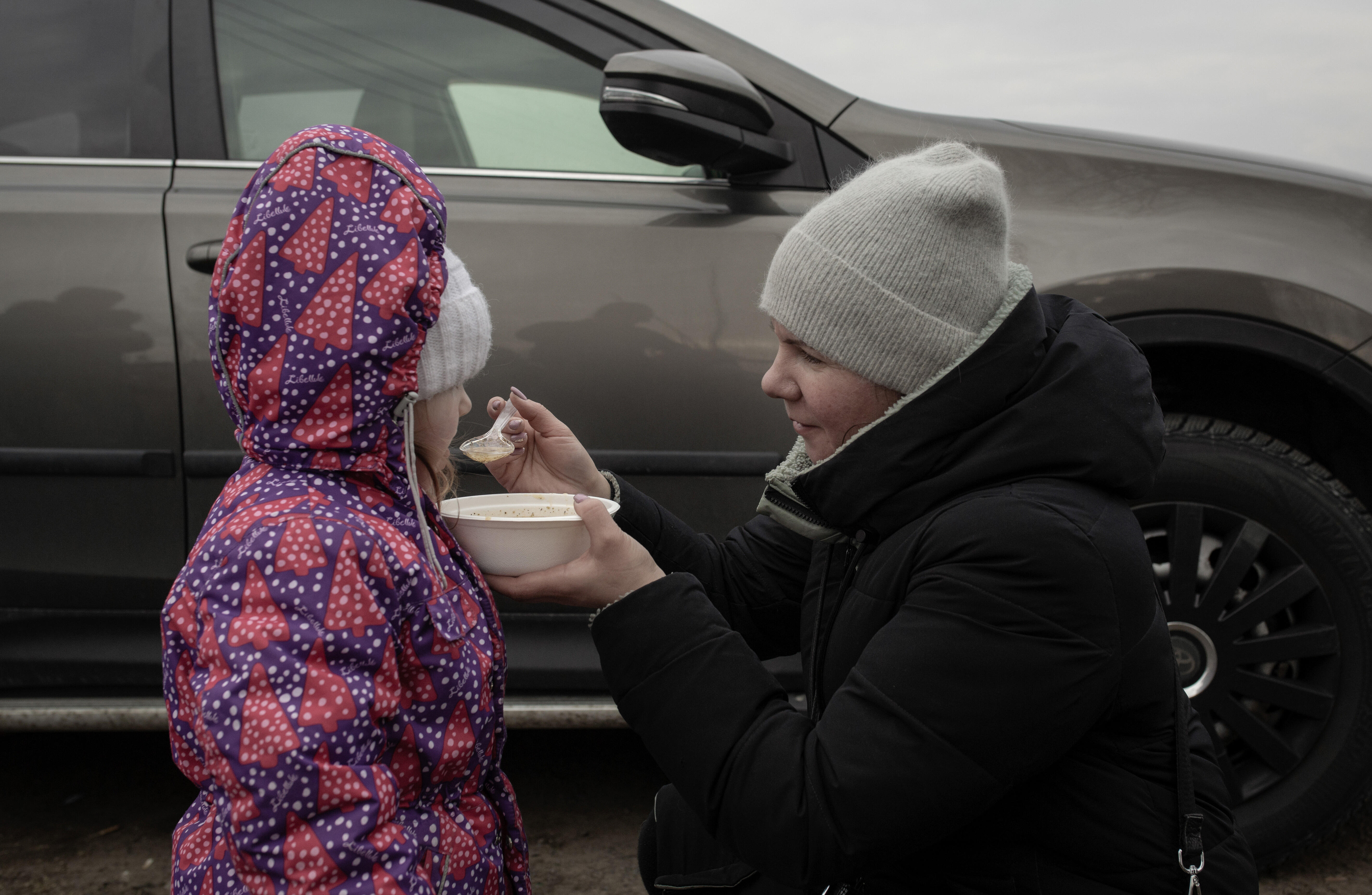 Dressed in winter clothes, a mother kneels down outside to feed her young daughter. 