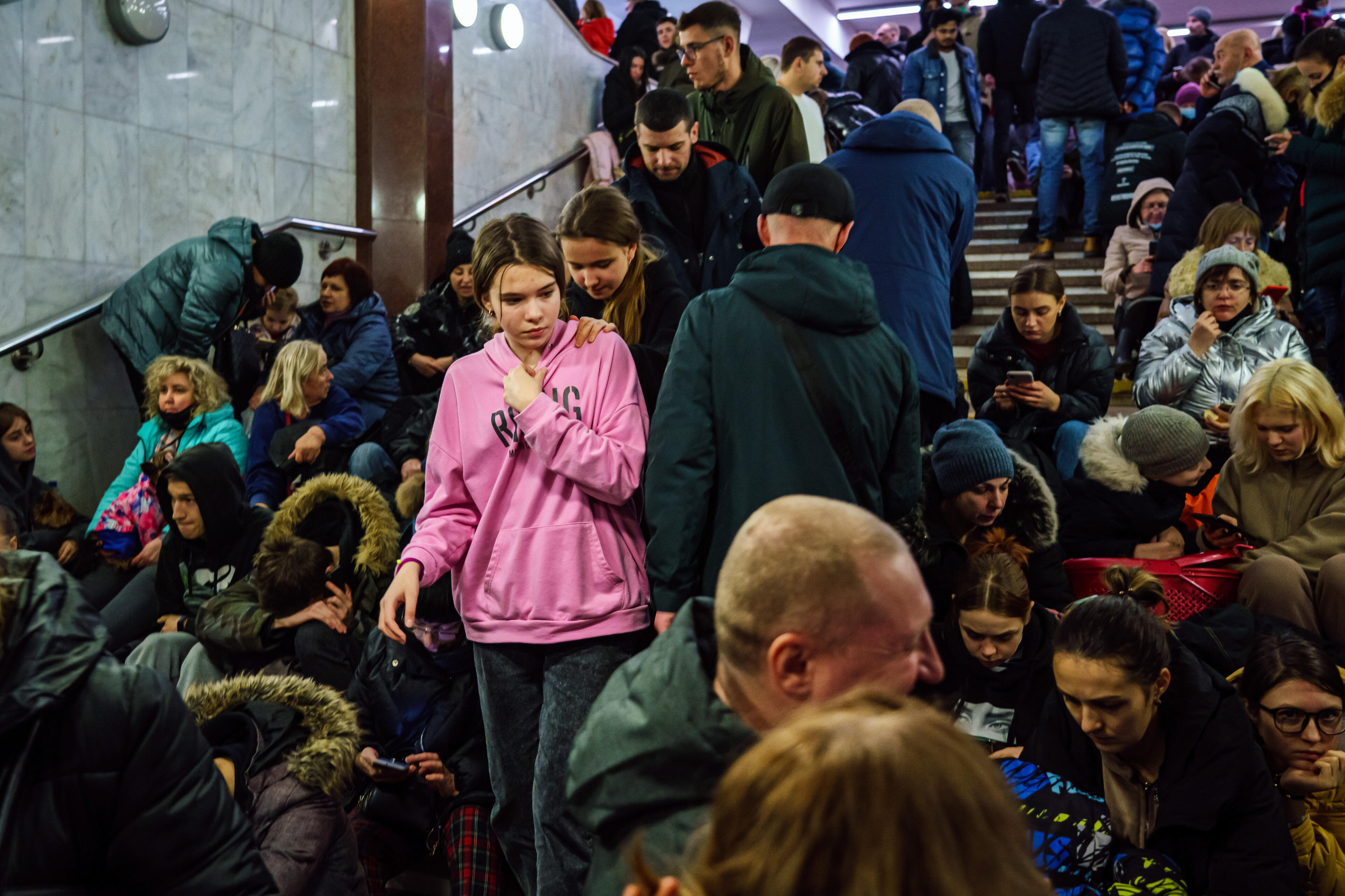 A girl wearing a hoodies stands among people seated on steps as they take shelter in a subway station amid ongoing Russian attacks on cities in Ukraine