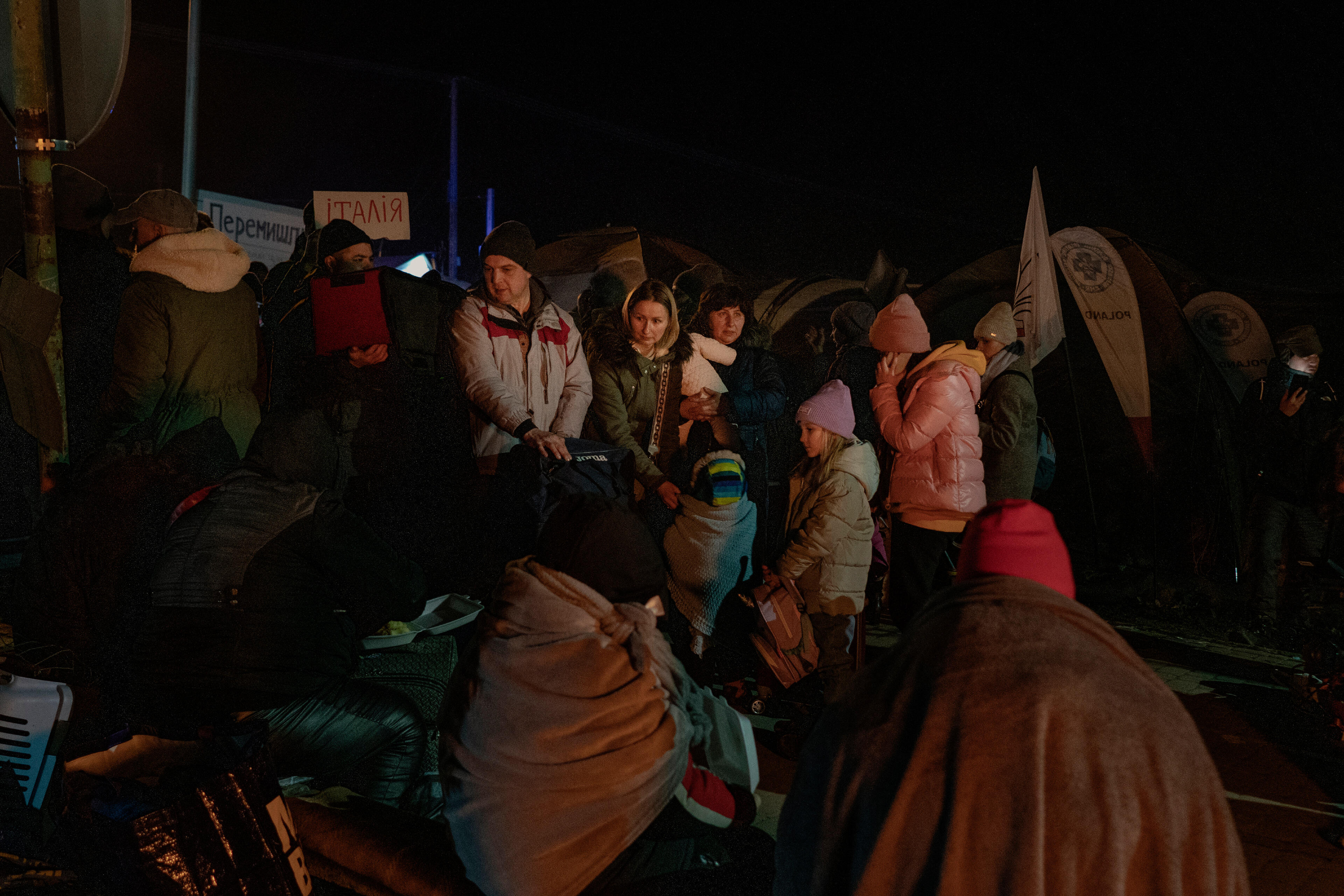 A small group of families with children who fled the war Ukraine, wearing winter coats, stand with their bags on a cold night at the Polish border at Medyka.