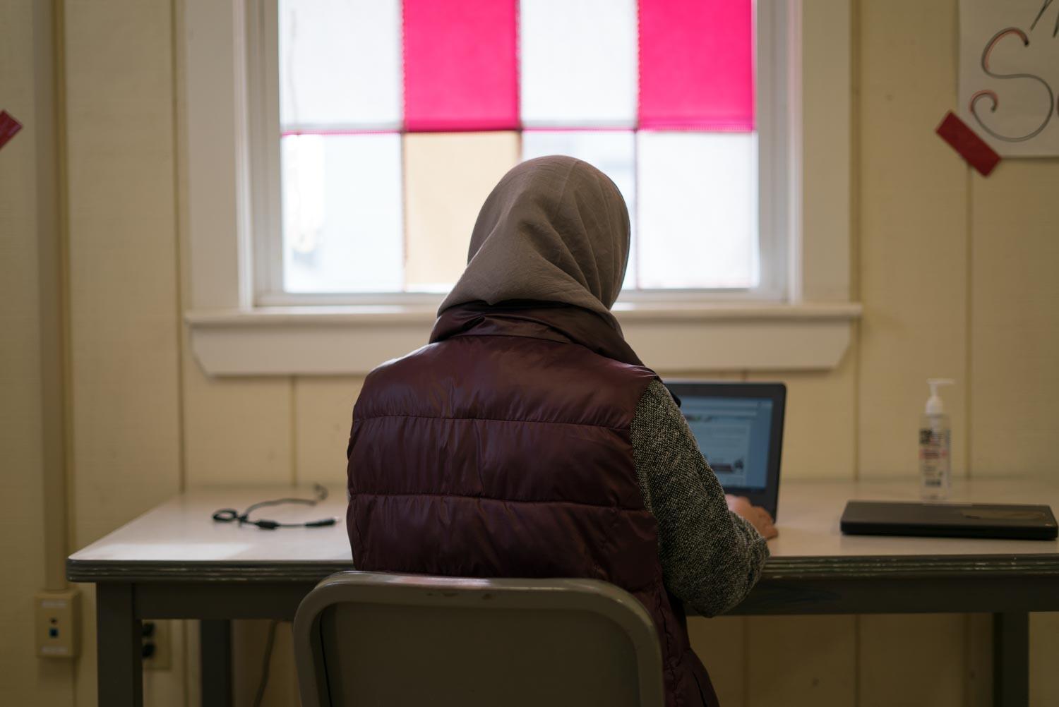 A young woman, with her back to the camera, sits at a desk and types on her laptop.