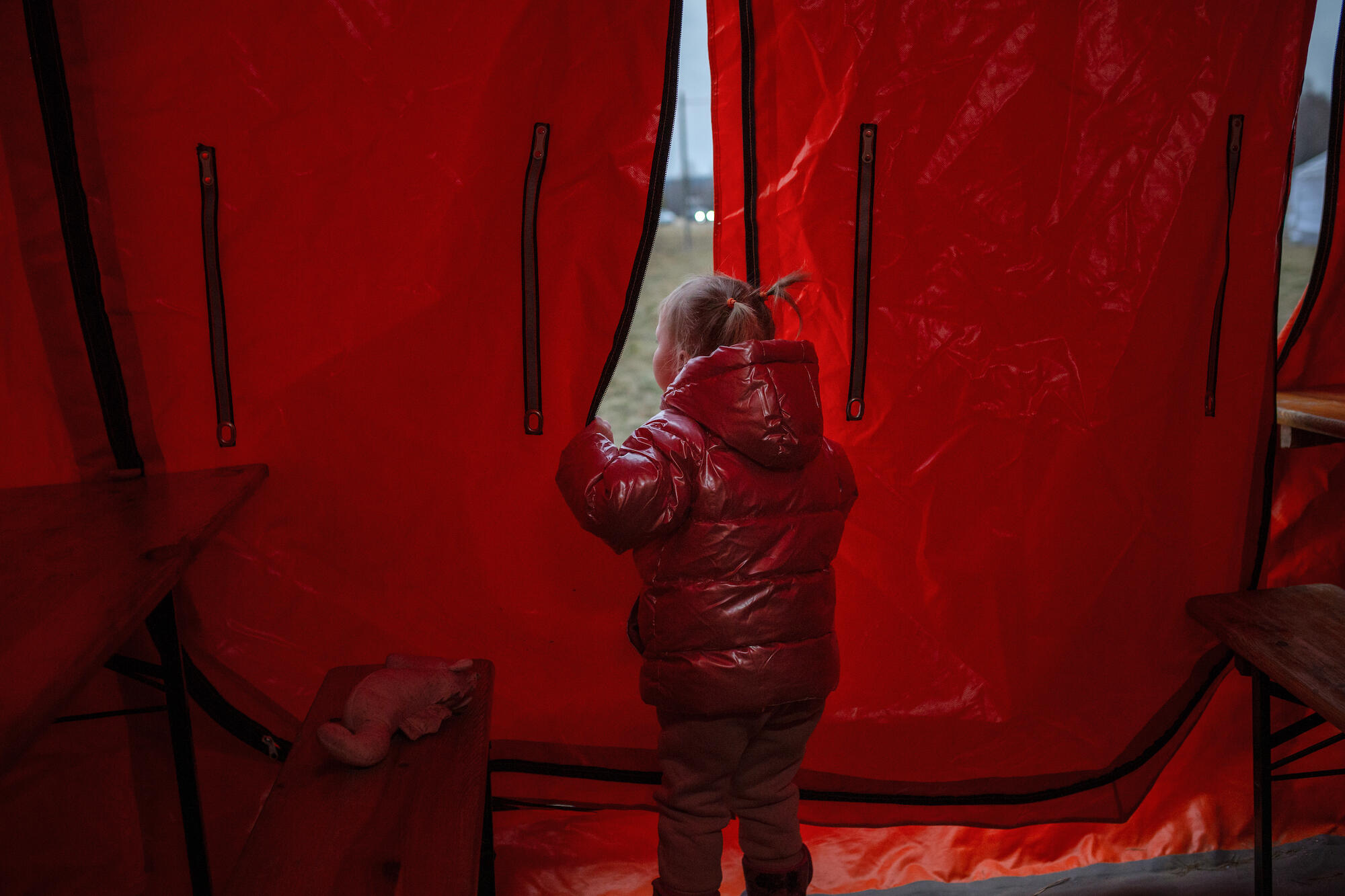 A young girl wearing a red winter coat stands in a red tent, peering out.