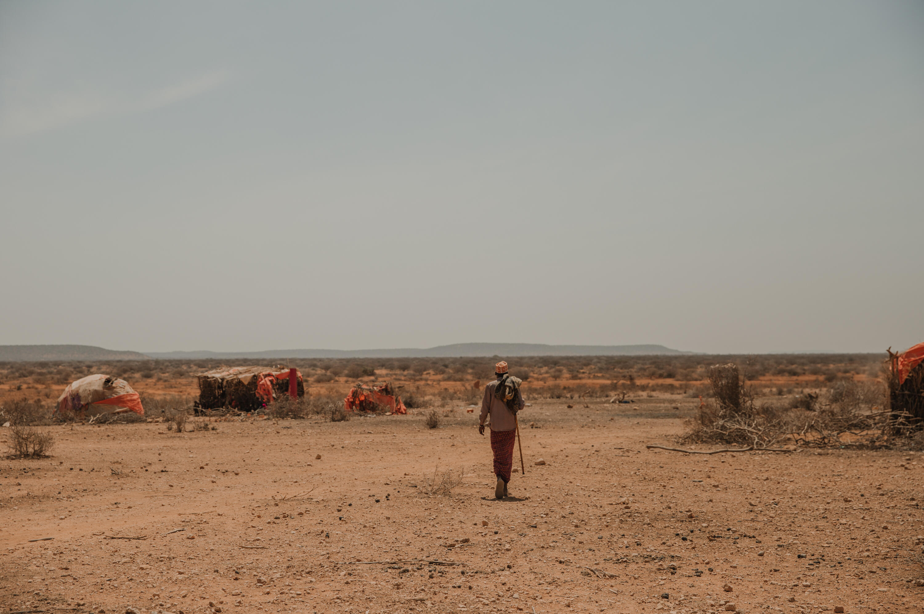 A man carrying a staff walks through a parched landscape in Ethiopia amid makeshift tents with mountains in the distance under a hazy sky.