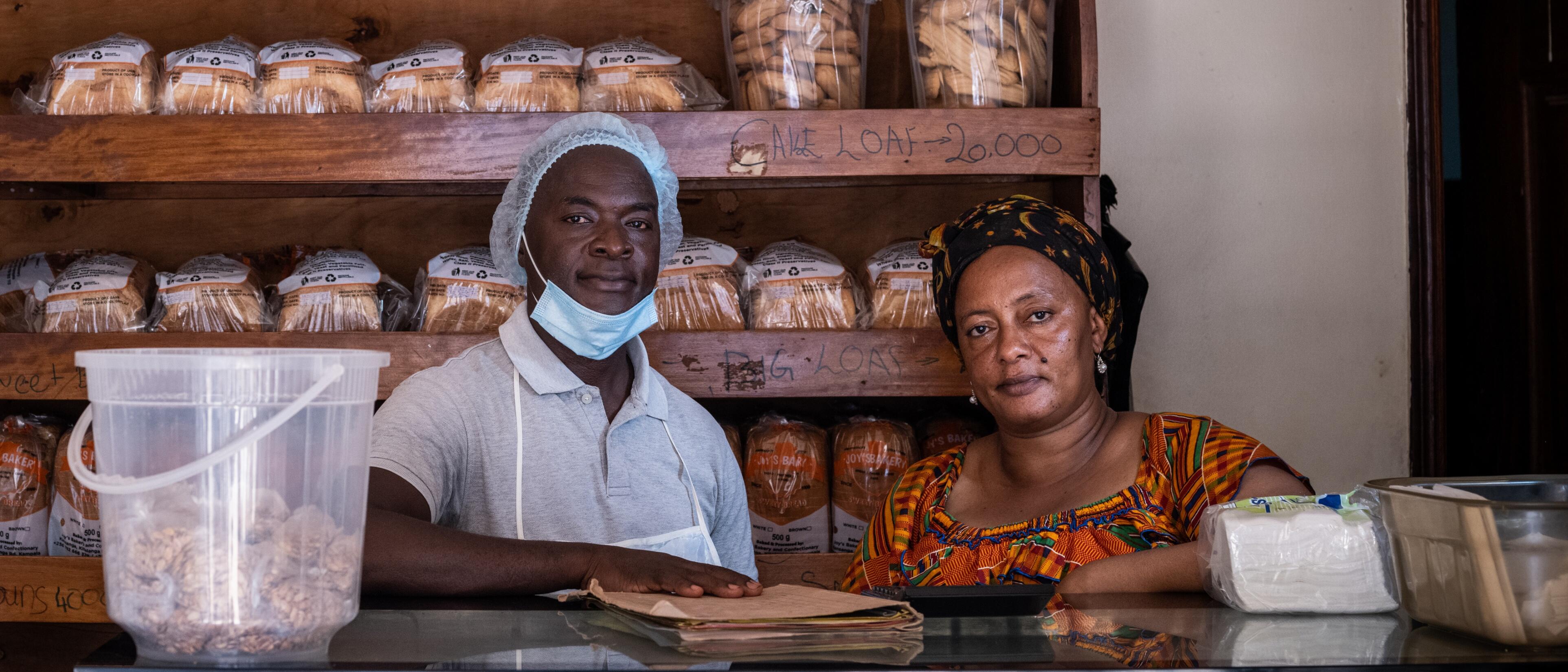 A Ugandan baker wearing a head covering, apron and lowered COVID face mask stands at his bakery counter with his mentee, a woman from South Sudan.