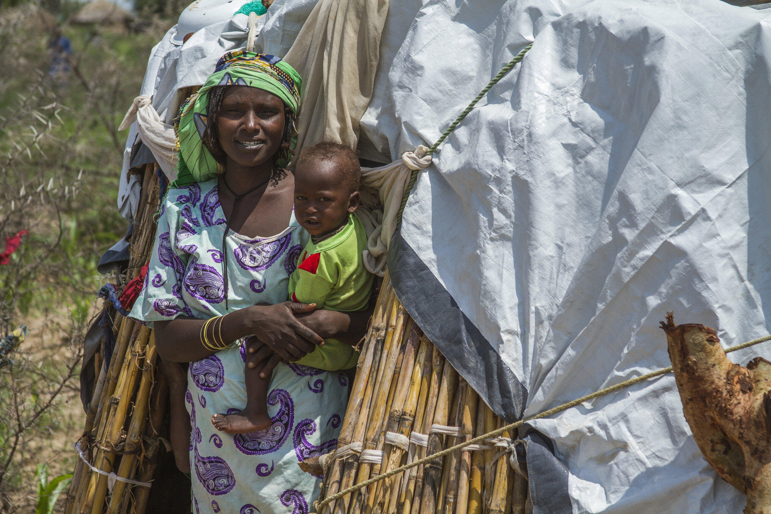 A displaced mother, smiling, holds her baby boy outside a makeshift tent in northern Cameroon.