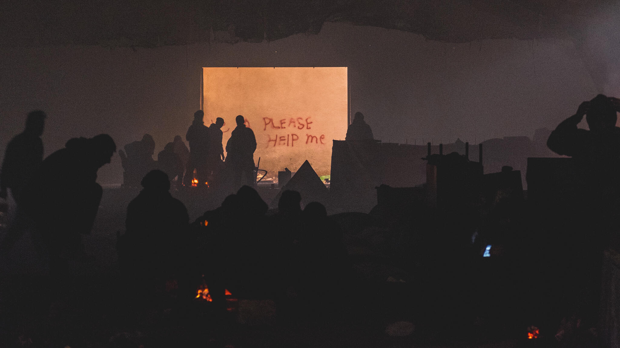 Refugees warm themselves by a bonfire at night on an abandoned industrial site in Belgrade, their figures silhouetted against a slab partly covered by graffiti that reads "PLEASE HELP ME." 
