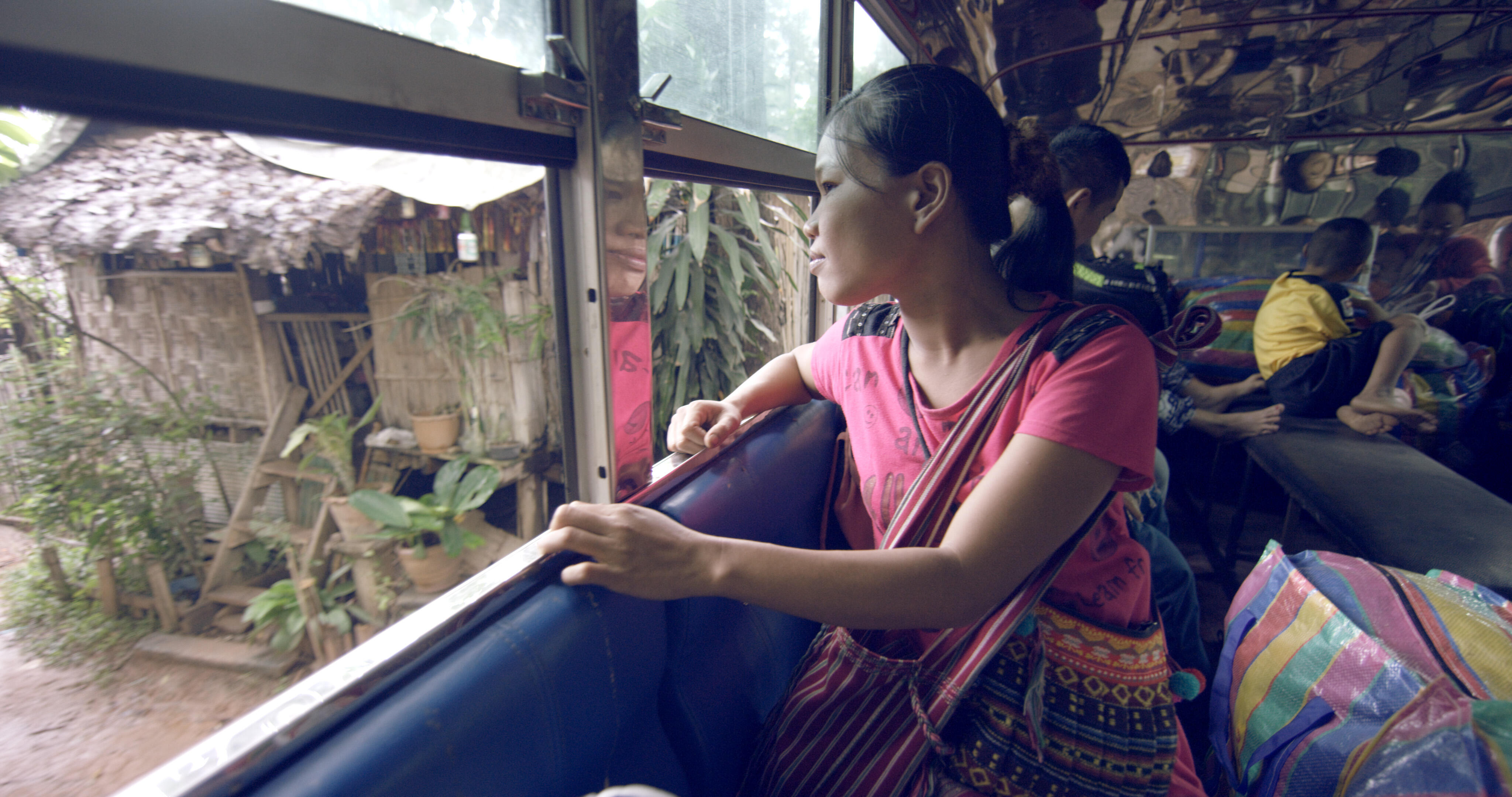 A young woman leaves a refugee camp in Thailand, looking out a bus window.