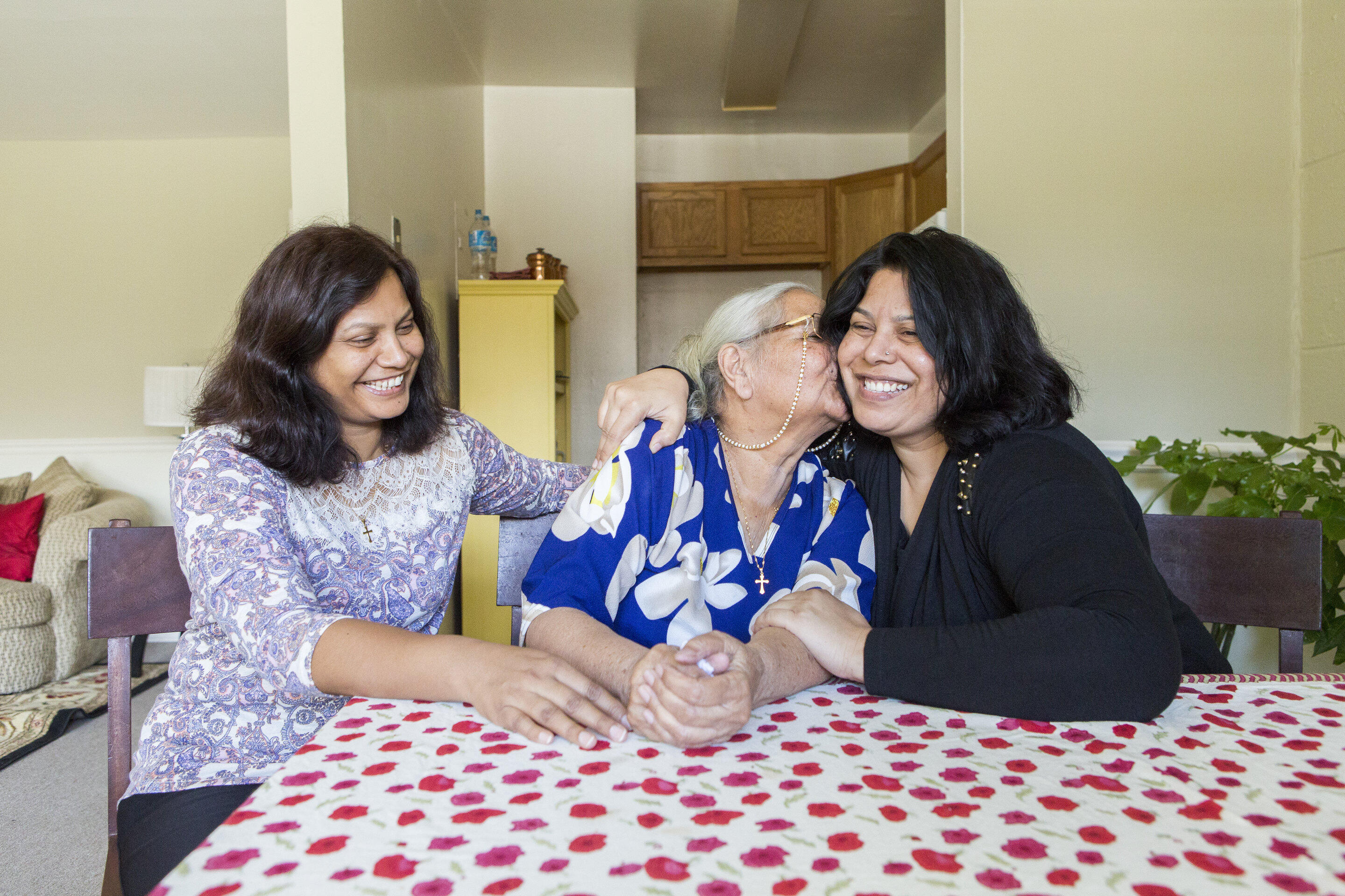 Three women, one older and two middle aged, sit at a table with a red and white table clothe. They are smiling and the older woman is kissing the woman on her left. They are all refugees. 