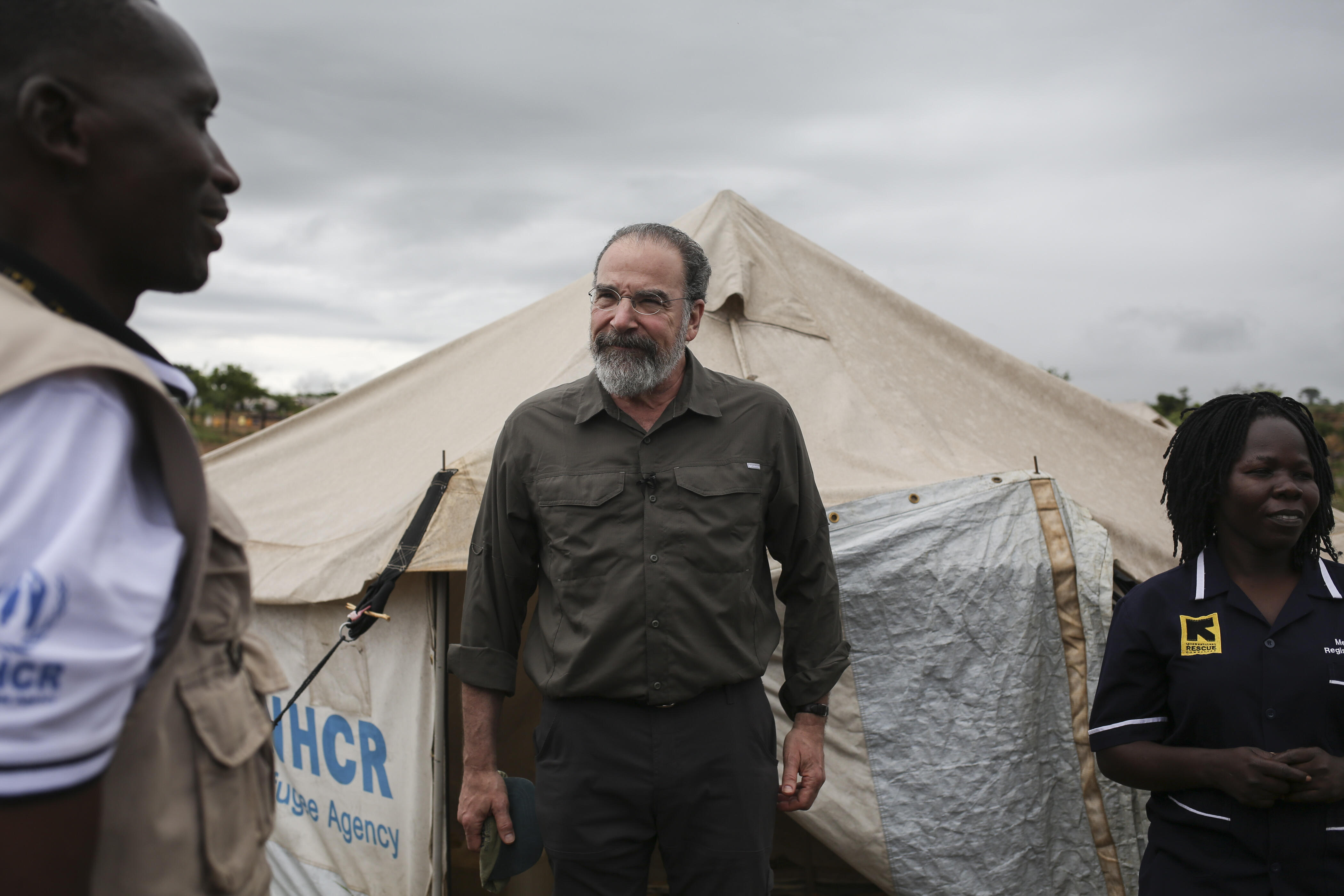 Mandy Patinkin with two IRC aid workers outside a tent in a refugee settlement in Uganda