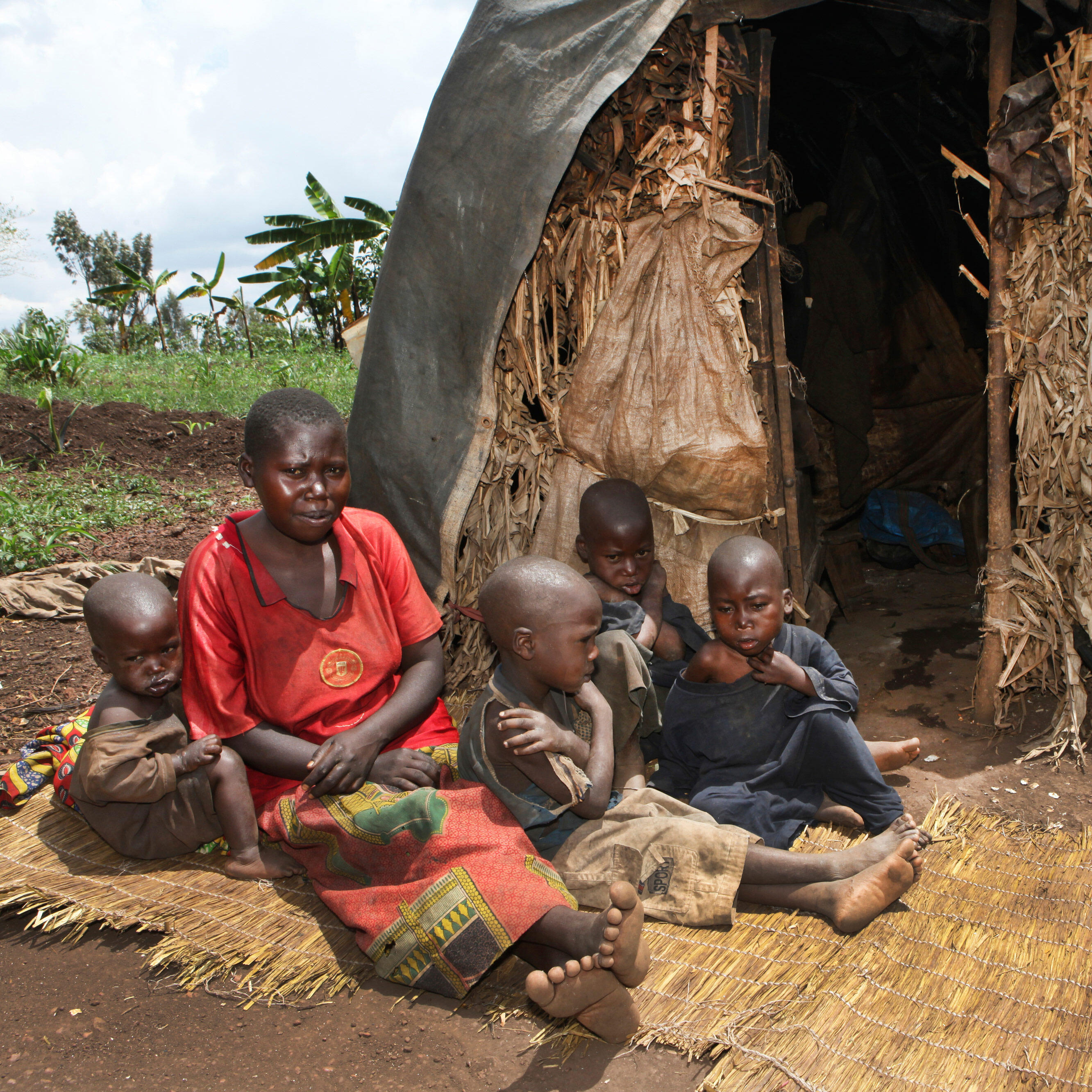 A Burundian woman and four children sit on straw mats on the the ground outside of their thatched home.