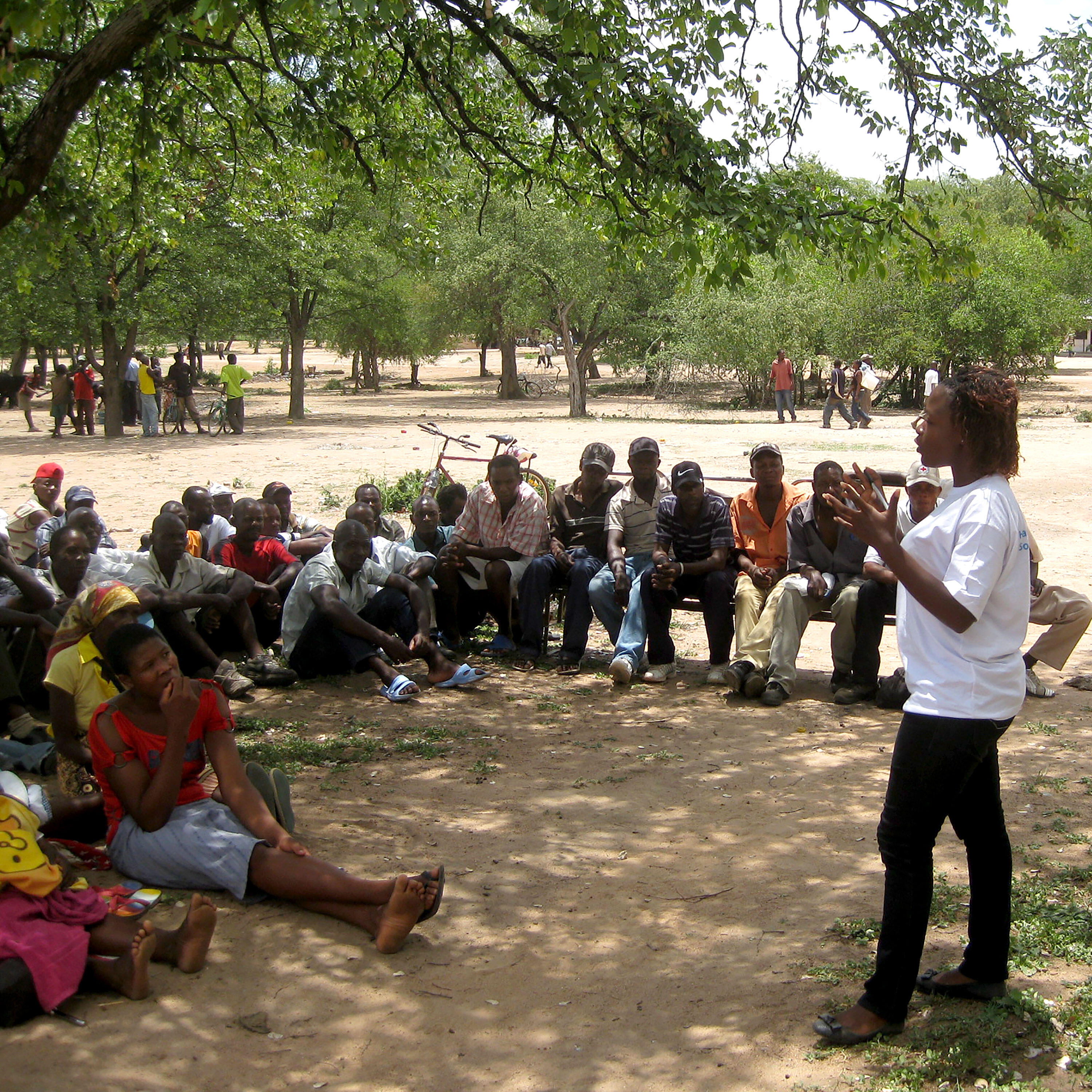 A woman who is an IRC staff member stands speaking animatedly to a group of people seated on the ground in the shade of a tree in Zimbabwe