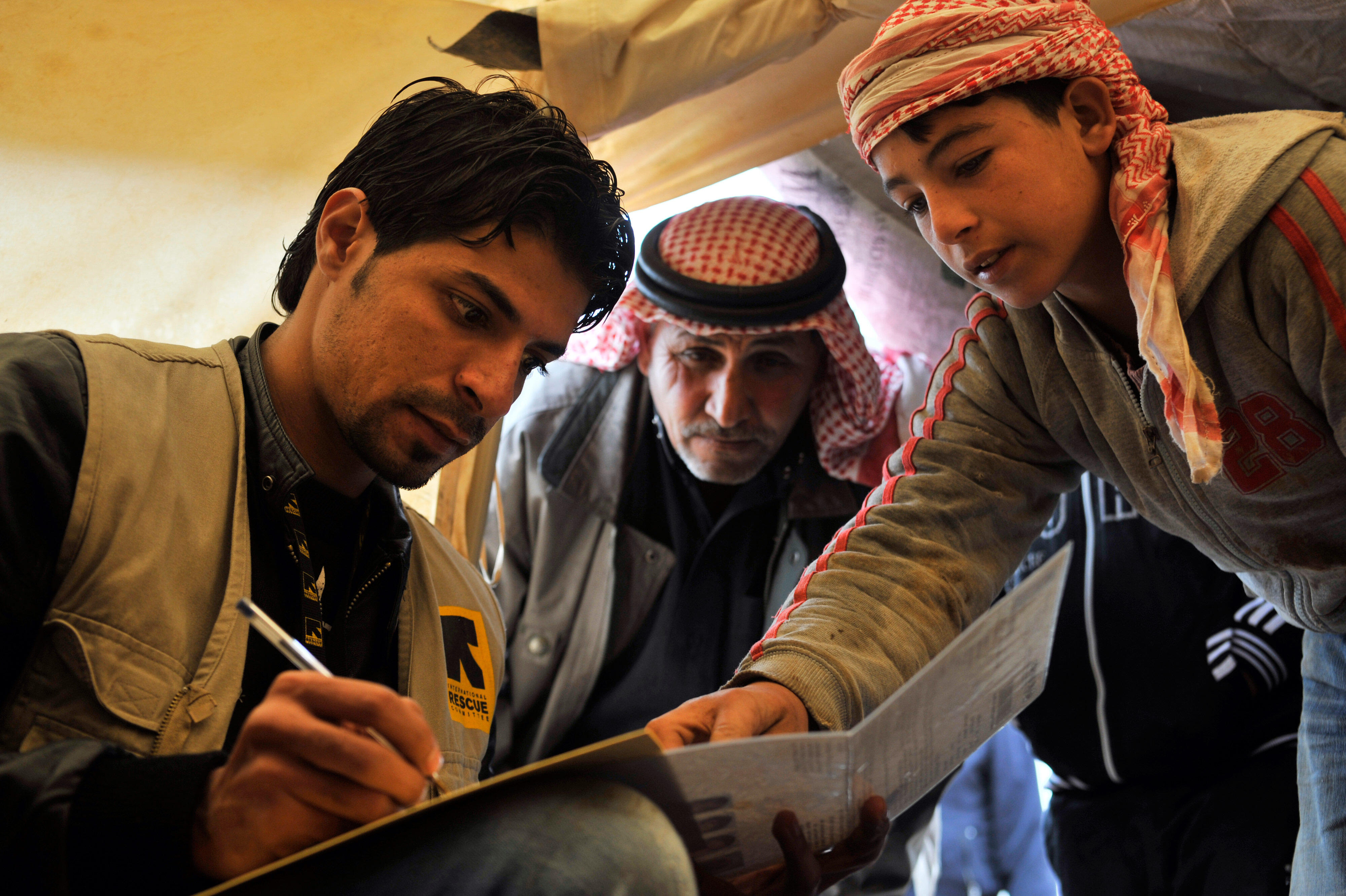 A volunteer from the International Rescue Committee holds a pen while two Syrian refugees point to an item on the page the volunteer is writing on.
