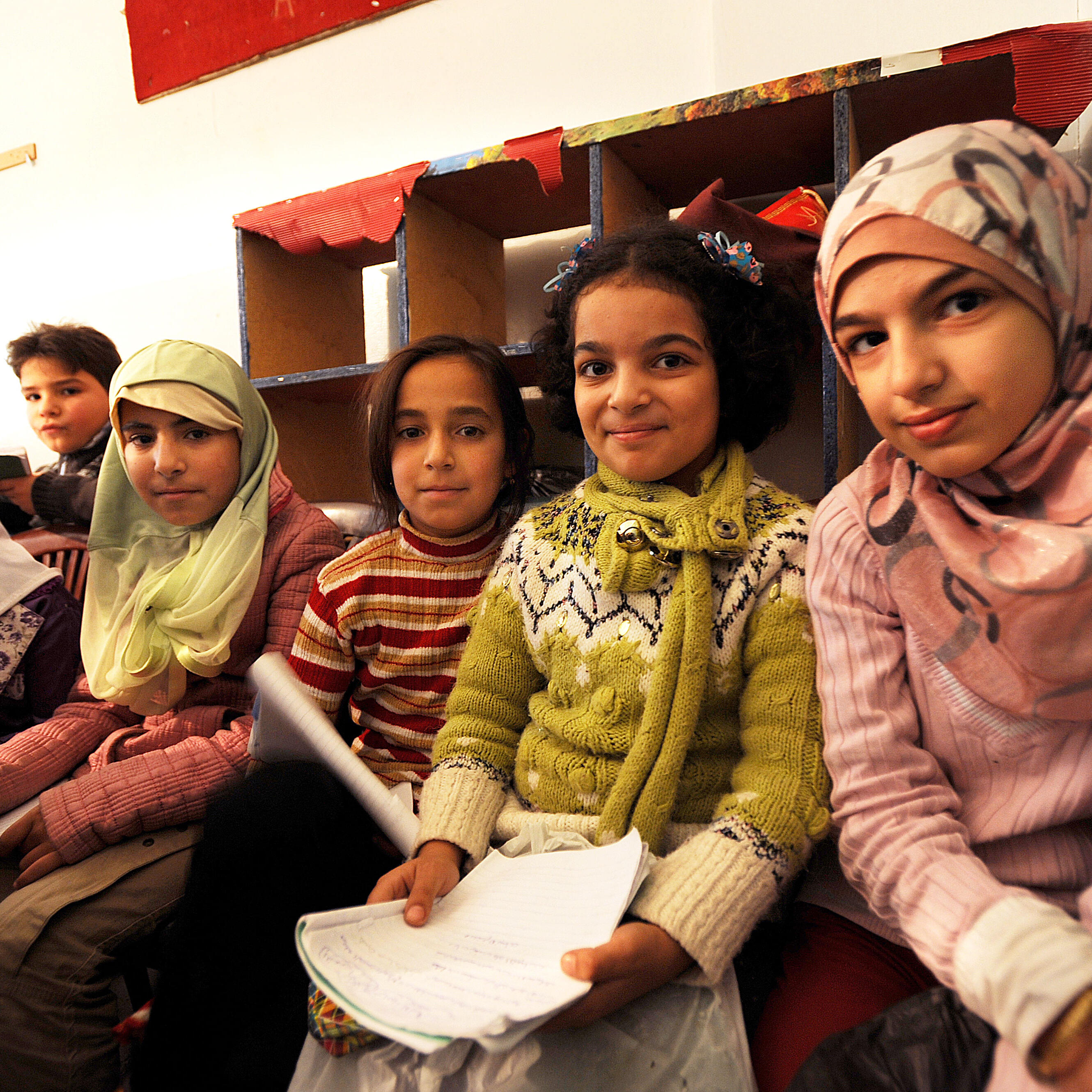 Five Syrian girls sitting in a school in Lebanon holding papers and pencils.