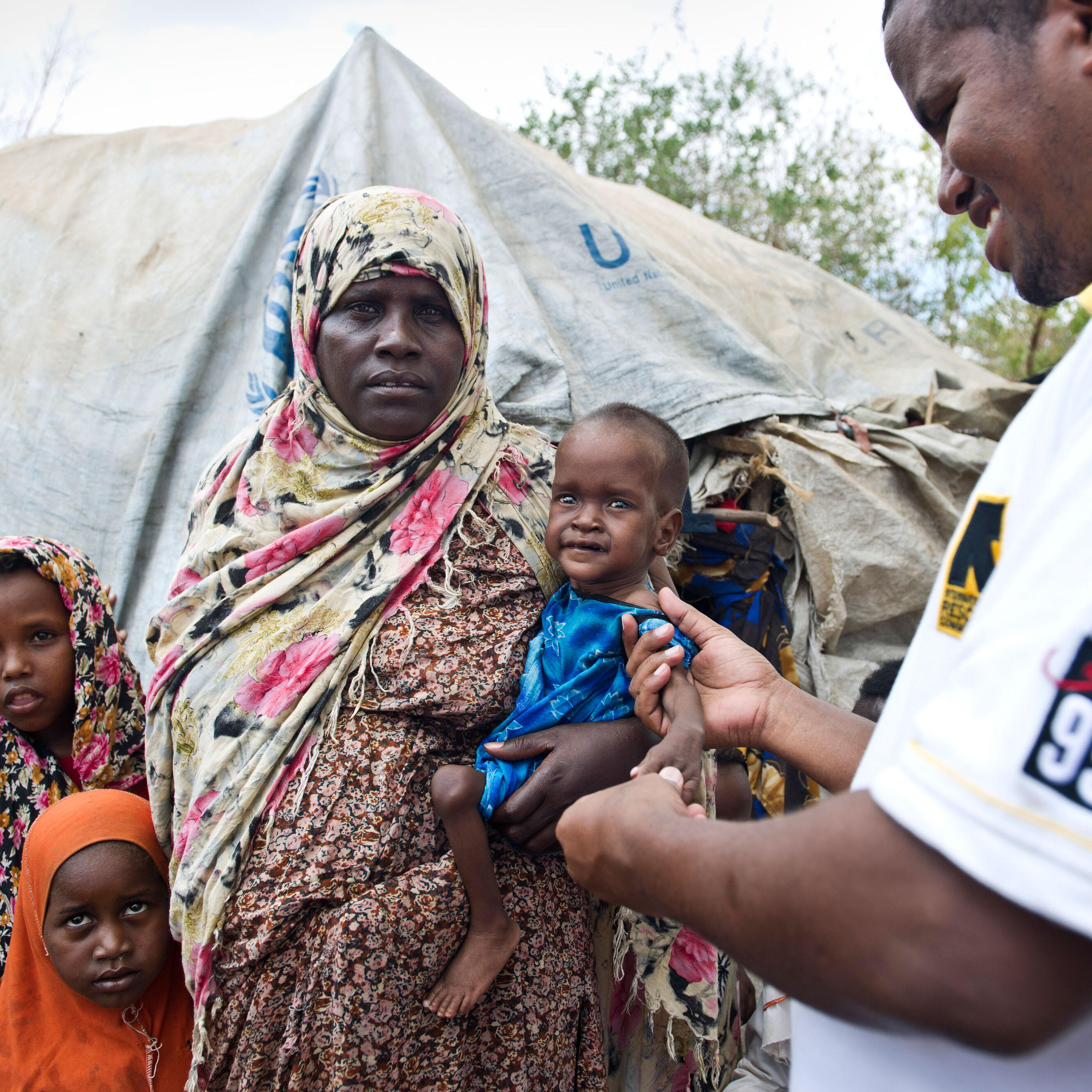 A male IRC health worker in Dadaab refugee camp stands outside a tent examining a baby girl being held in her mother's arms for signs of malnutrition.