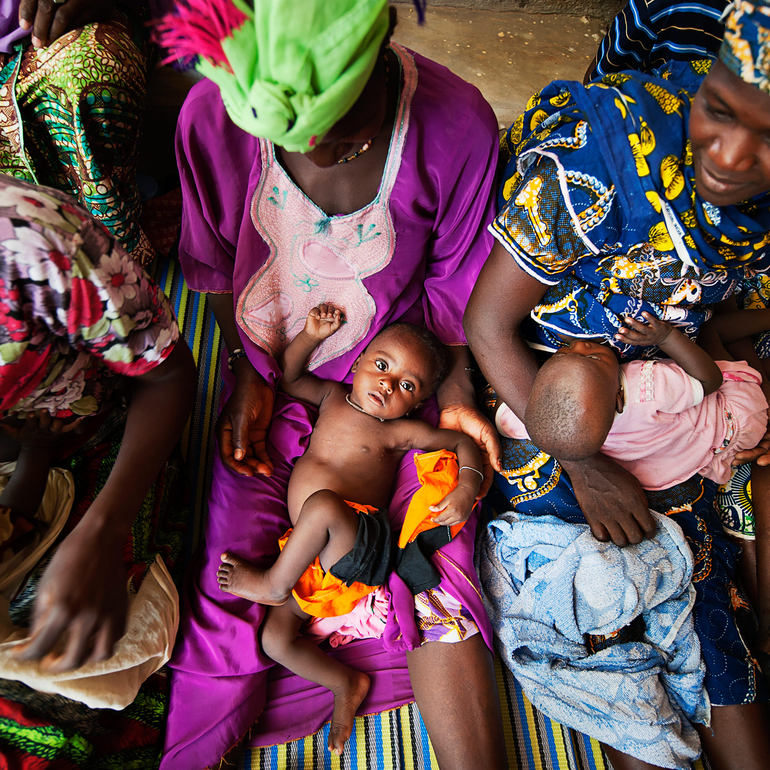 Malian women wearing brightly colored clothing hold children as they are measured and weighed.