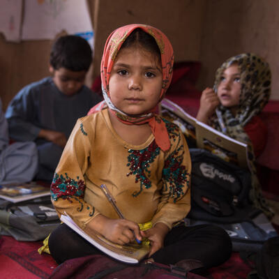Five children sit on the ground in a school with books and school supplies surrounding them. One girl sits in the foreground on the photo and looks at the camera while holding a notebook. 