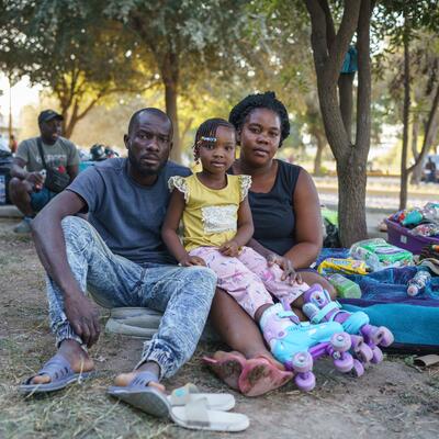 In a makeshift encampment in Mexico, a Haitian family--a mom, dad and young daughter--look straight at the camera while sitting on the ground next to their suitcases and blankets. 