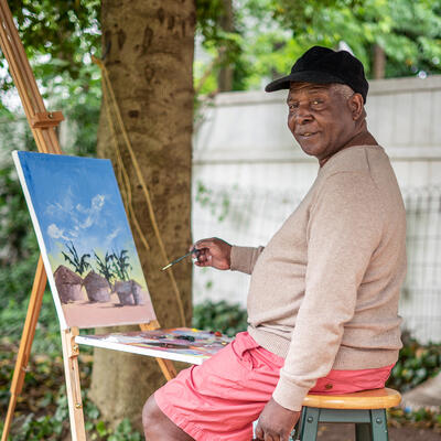 Muyambo Marcel Chishimba is a painter and recently resettled refugee from the Democratic Republic of Congo Chishimba sits on a stool in front a painting on an easel outside.